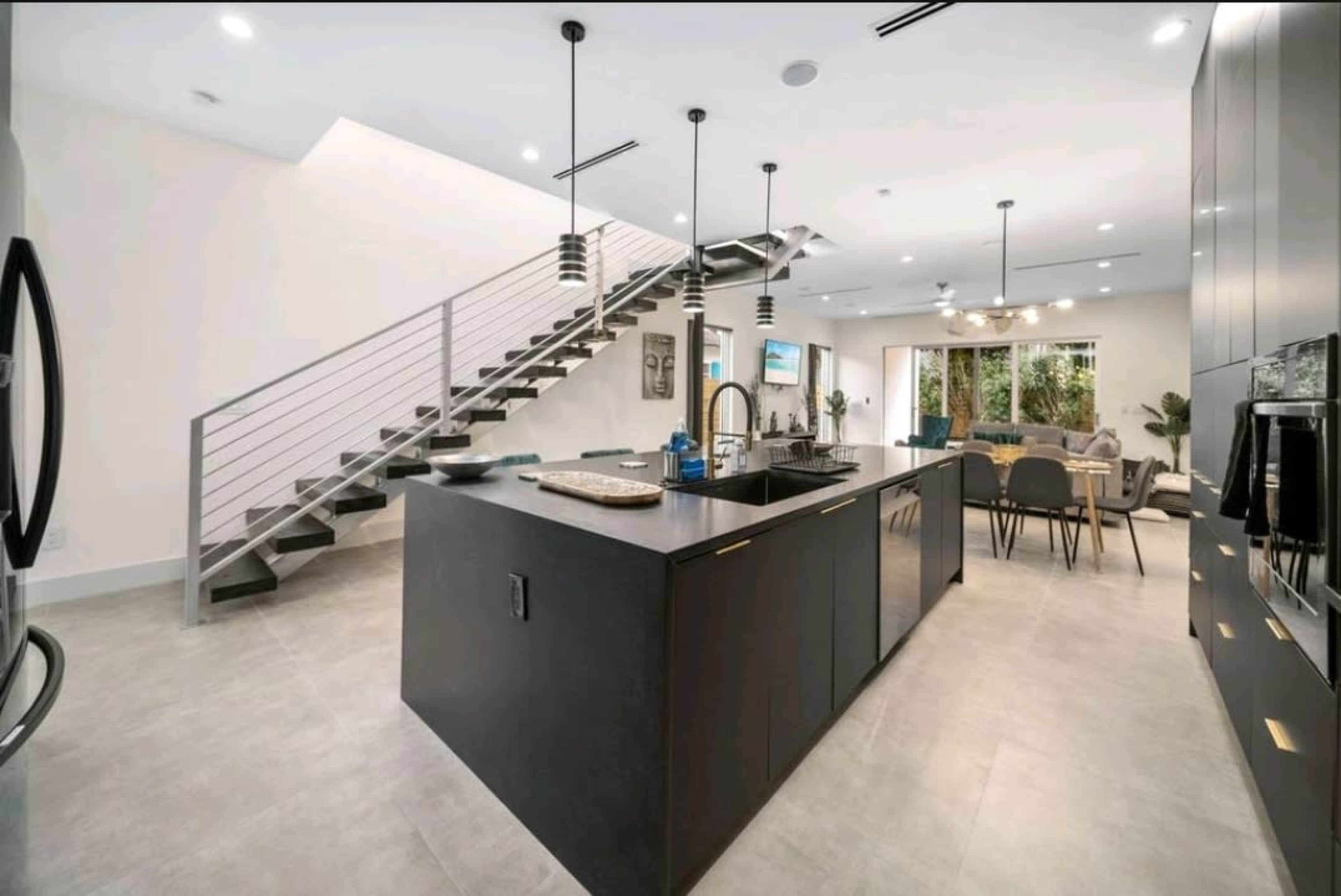 A modern kitchen features a large island with a sink, black cabinetry, and a staircase in the background.