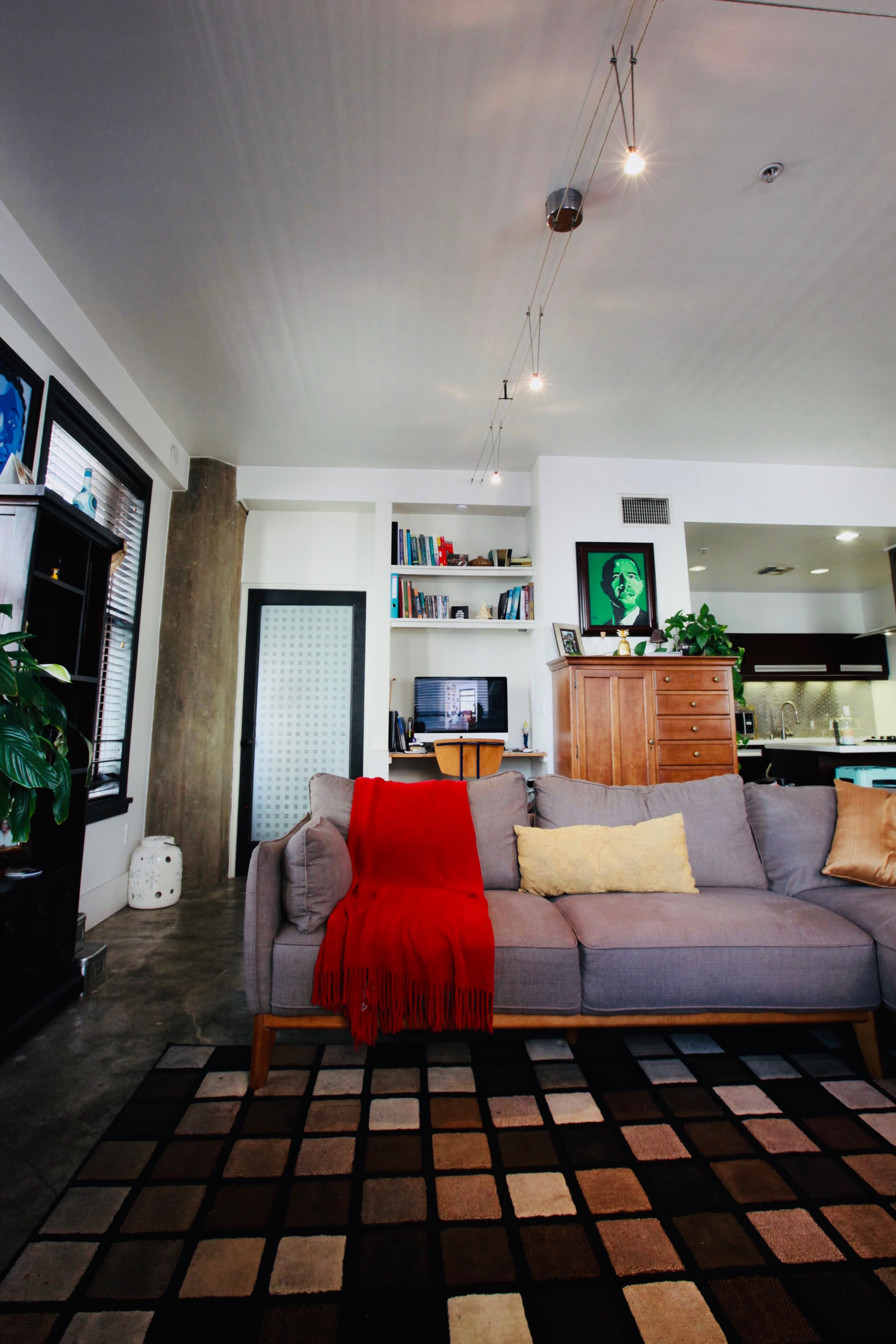 A modern living room features a gray sofa with a red throw blanket, a patterned rug, and built-in shelves displaying books and decorative items.