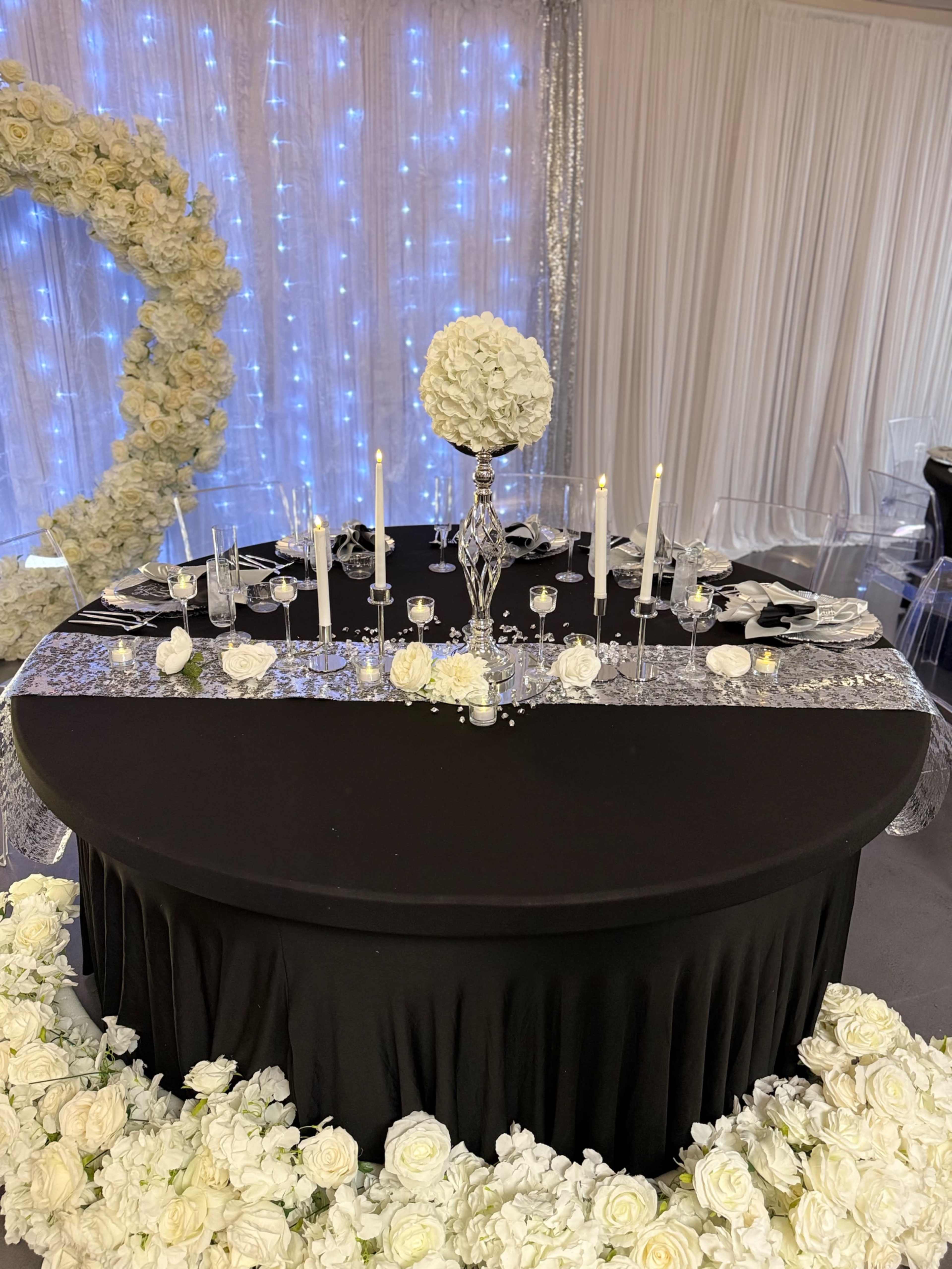 A round black table is elegantly set with white flower arrangements, candles, and glassware, surrounded by white floral decorations.