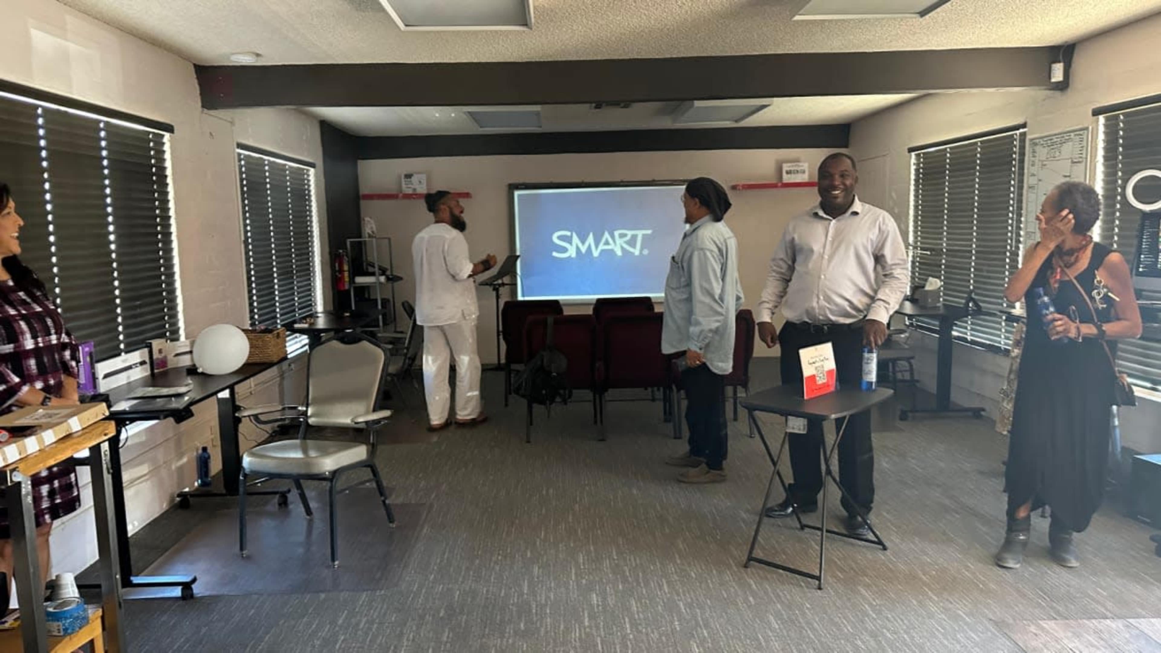 The image shows a small meeting room with chairs arranged in front of a projector screen displaying the word "SMART," while individuals engage with one another in the space.