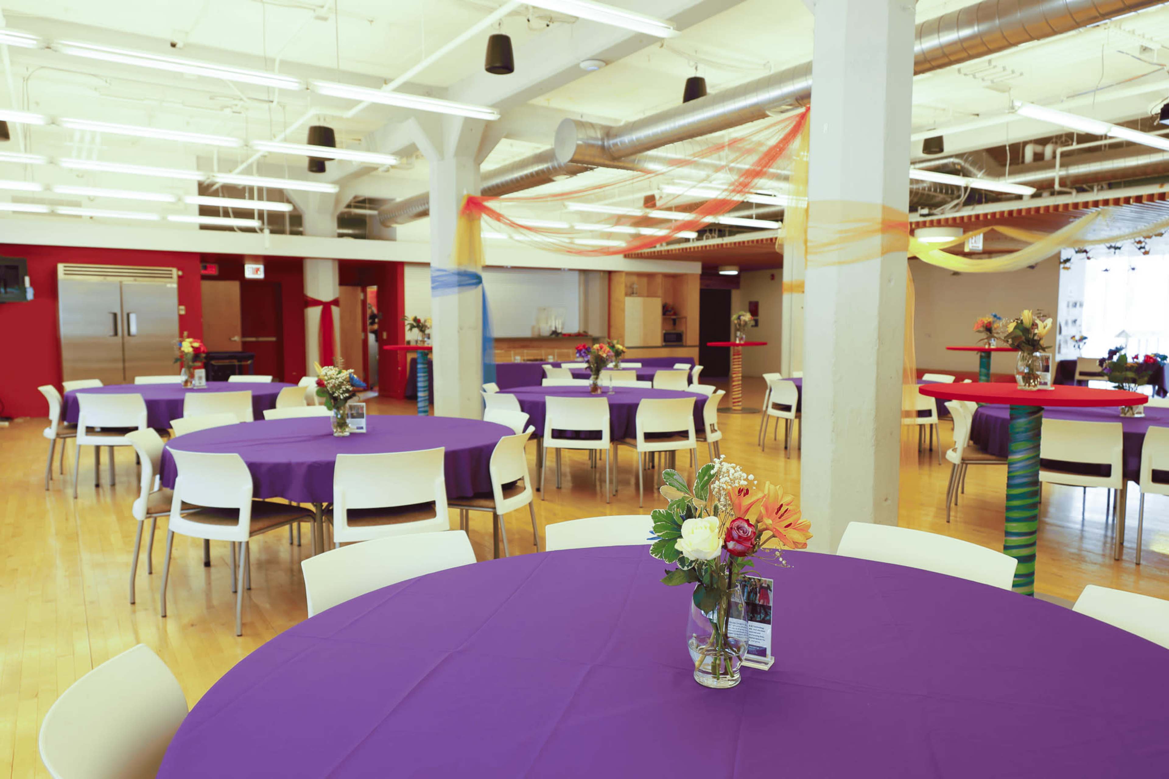 The image shows a spacious event room with several round tables covered in purple tablecloths, each adorned with vases of flowers, and colorful decorations hanging from the ceiling.
