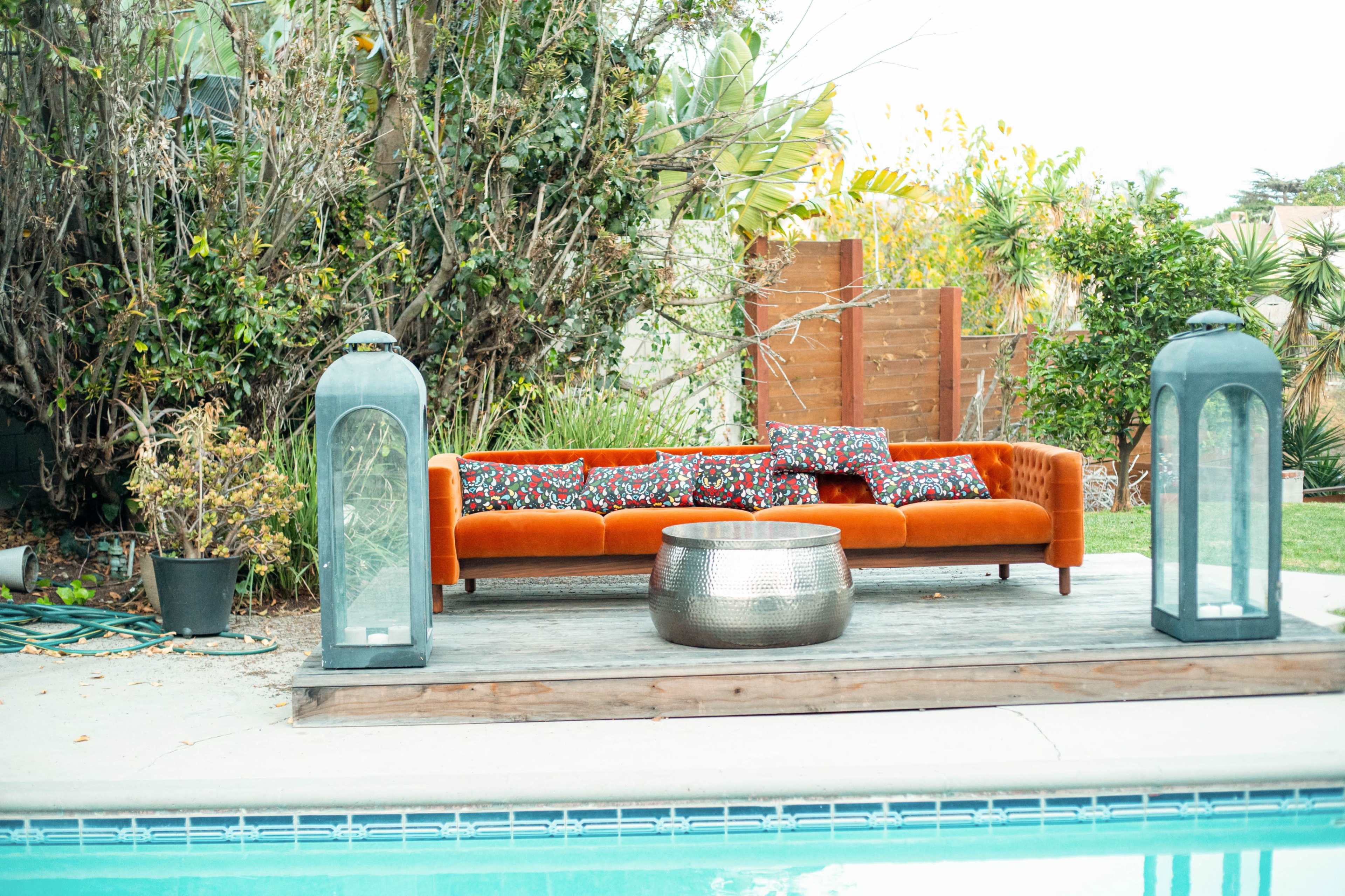 A bright orange couch with patterned pillows is placed on a wooden deck next to a pool, flanked by two large lanterns.