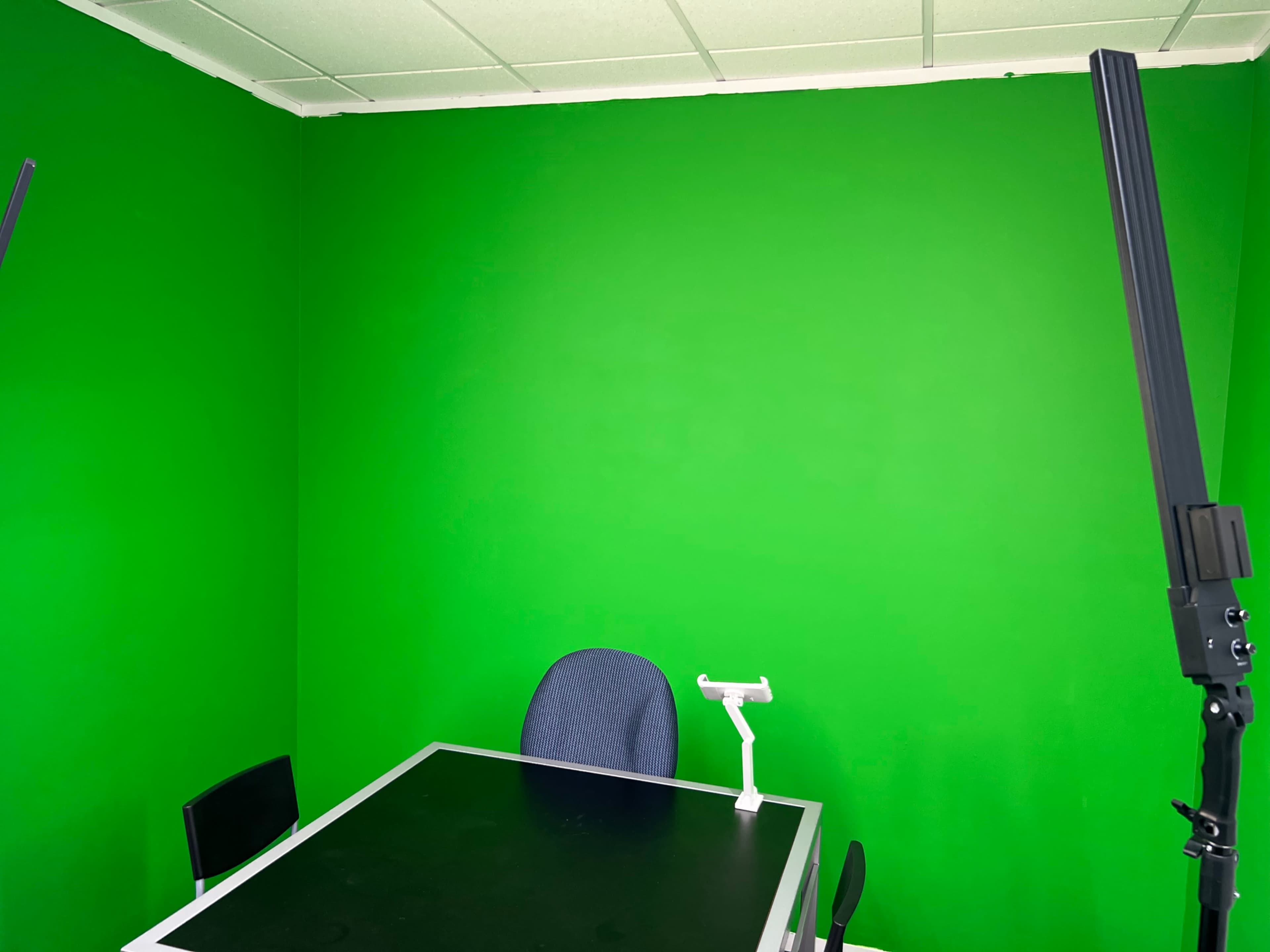 The image shows a small room with a green screen wall, a black table, and a single chair against the wall, with two lighting stands on either side.