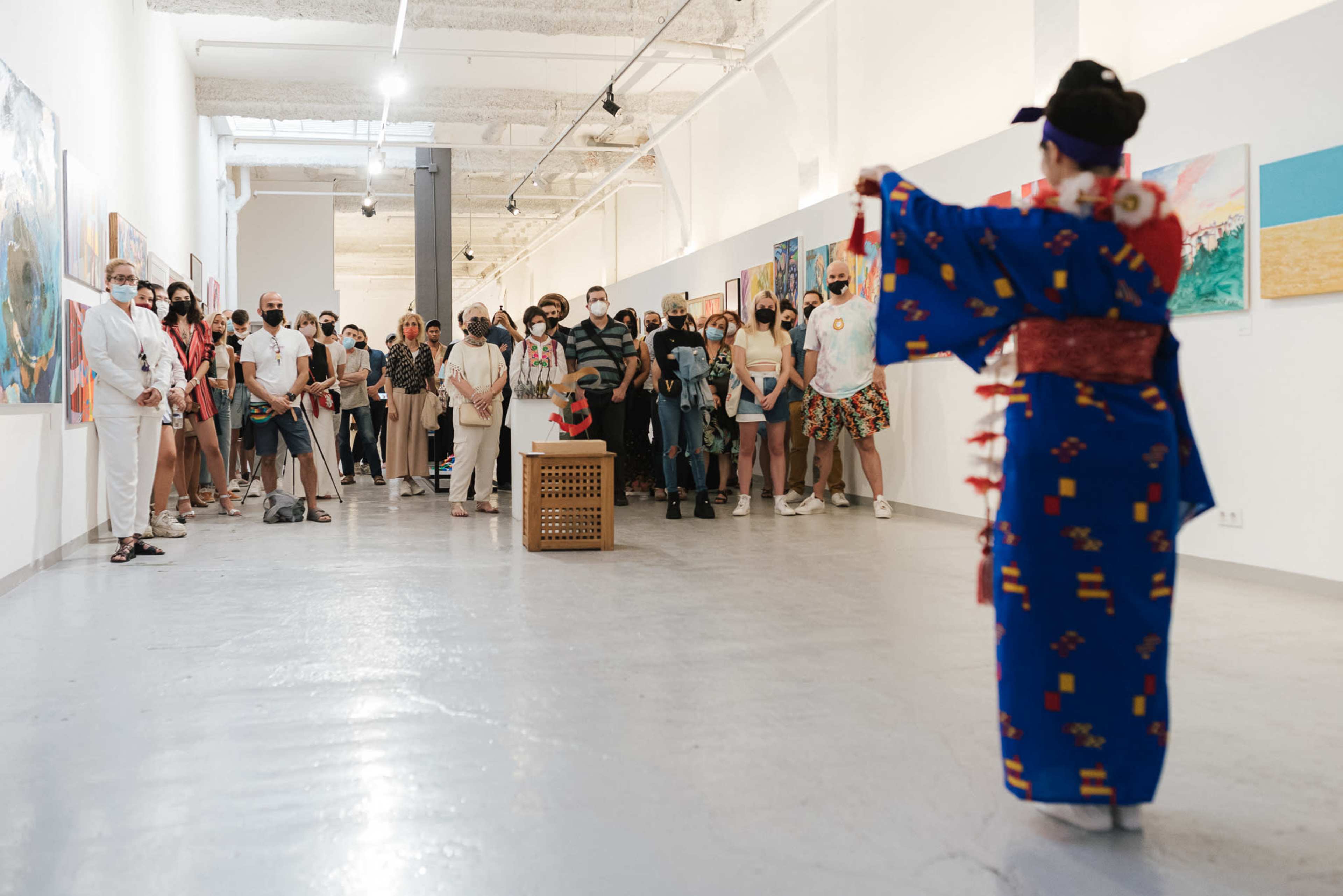 A group of people in masks watch a performer in traditional clothing at an art gallery event.