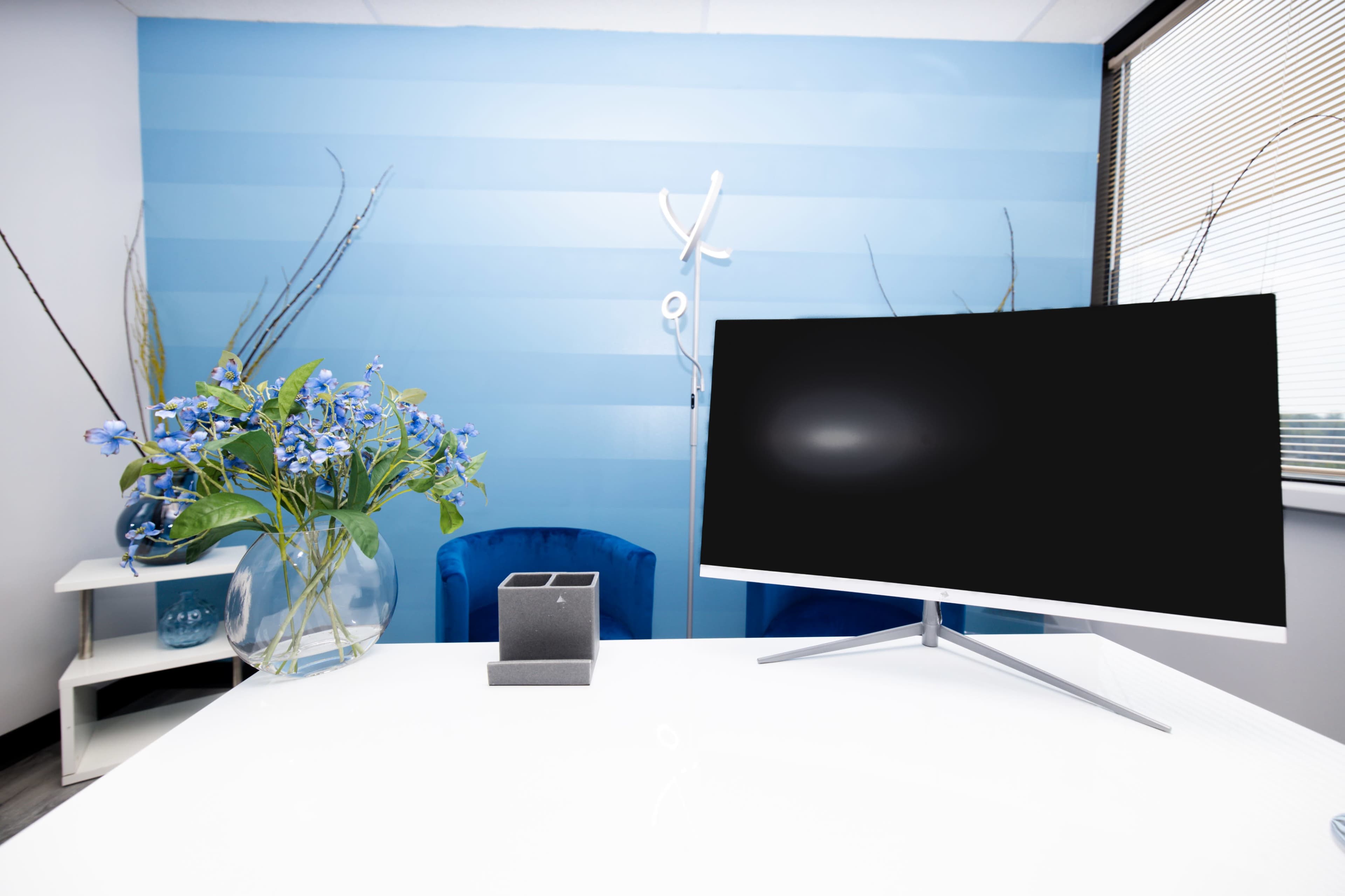 A modern office setting with a large computer monitor, a square pen holder, a vase of blue flowers, and a blue chair against a striped wall.