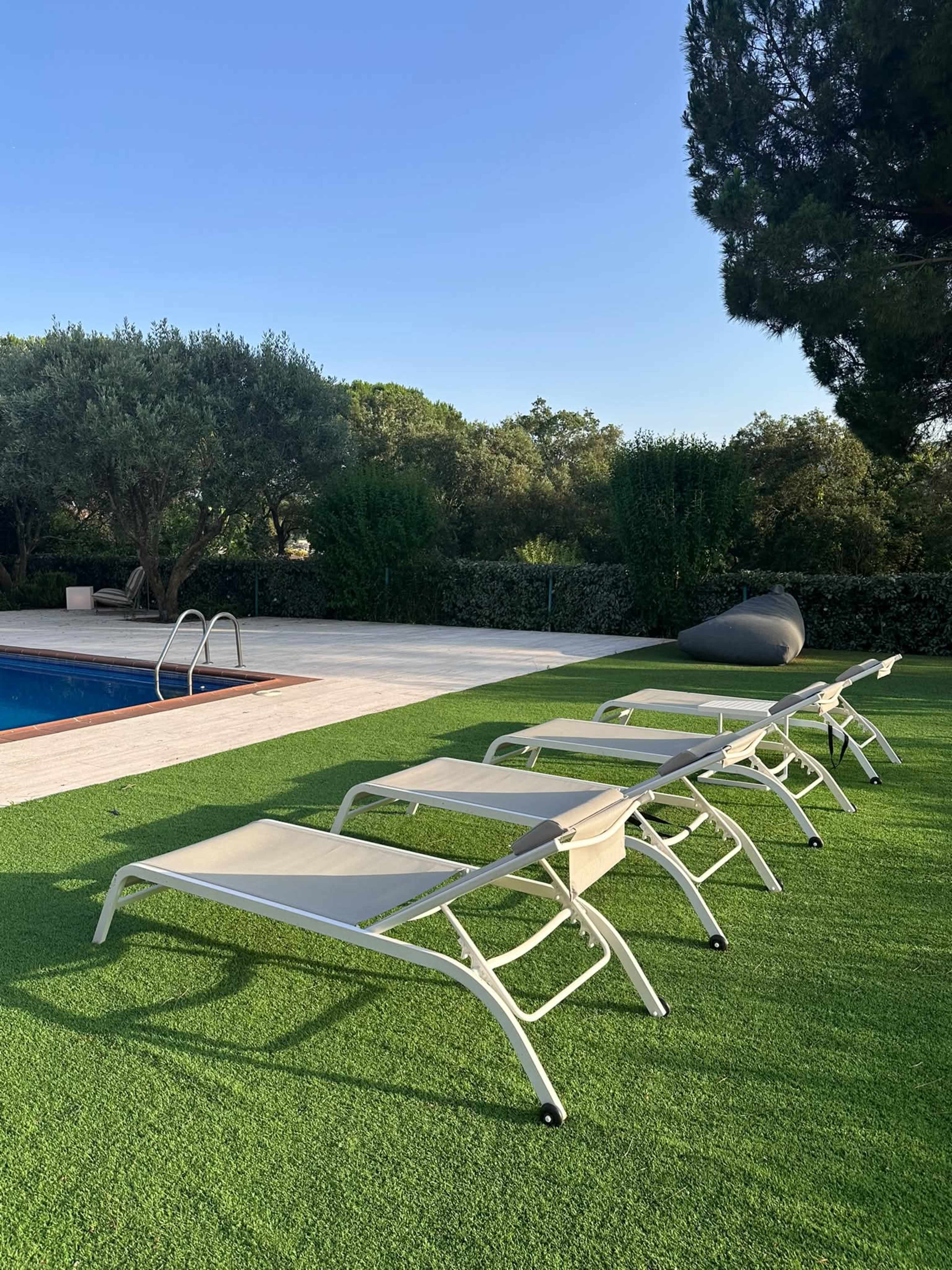 The image shows a row of five lounge chairs positioned on green artificial grass near a swimming pool surrounded by trees.