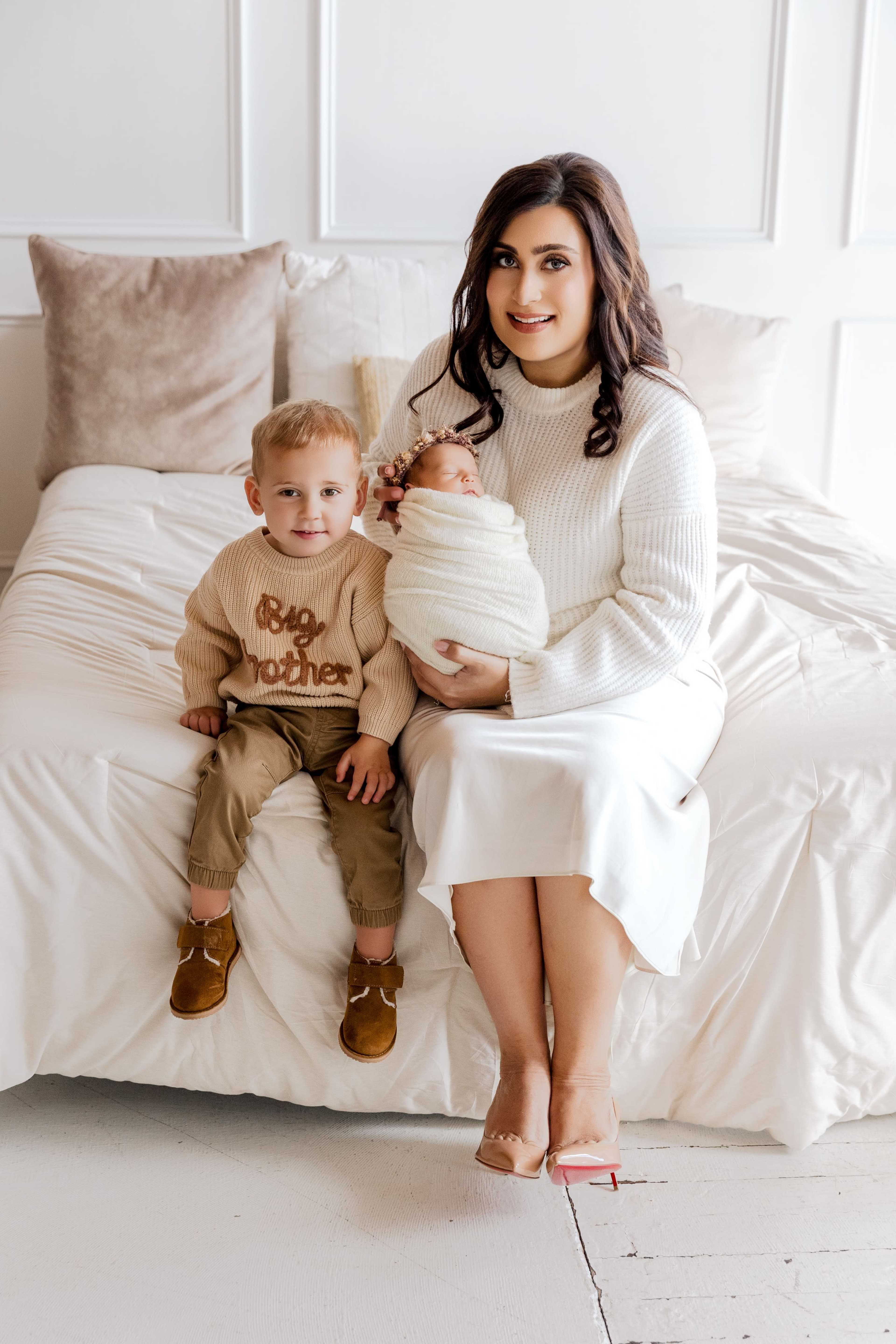 A woman sitting on a bed holds a newborn wrapped in a blanket, while a young boy sits next to her wearing a "Big Brother" sweater.