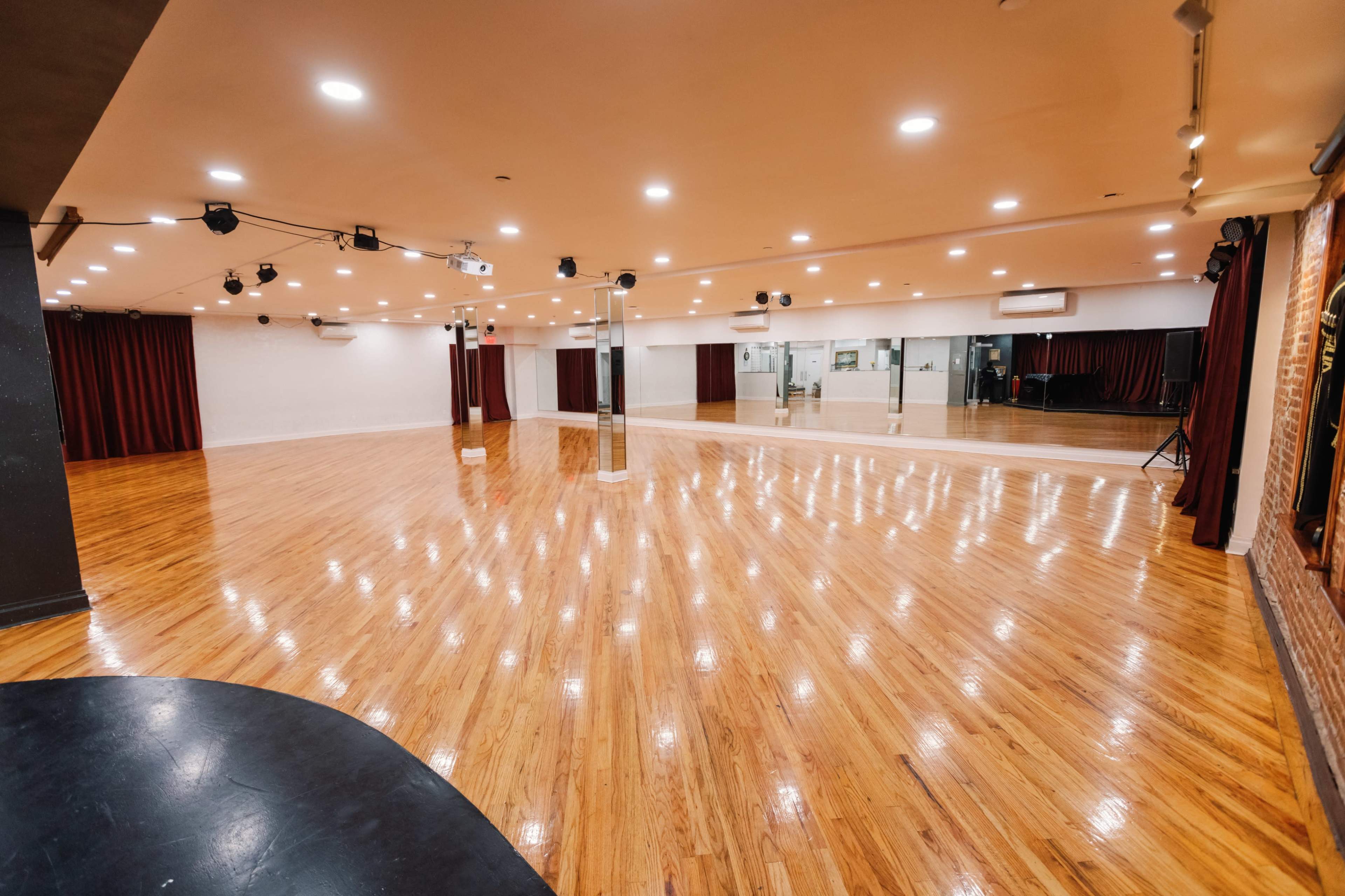 A spacious dance studio with polished wooden floors, large mirrors, and overhead lighting.