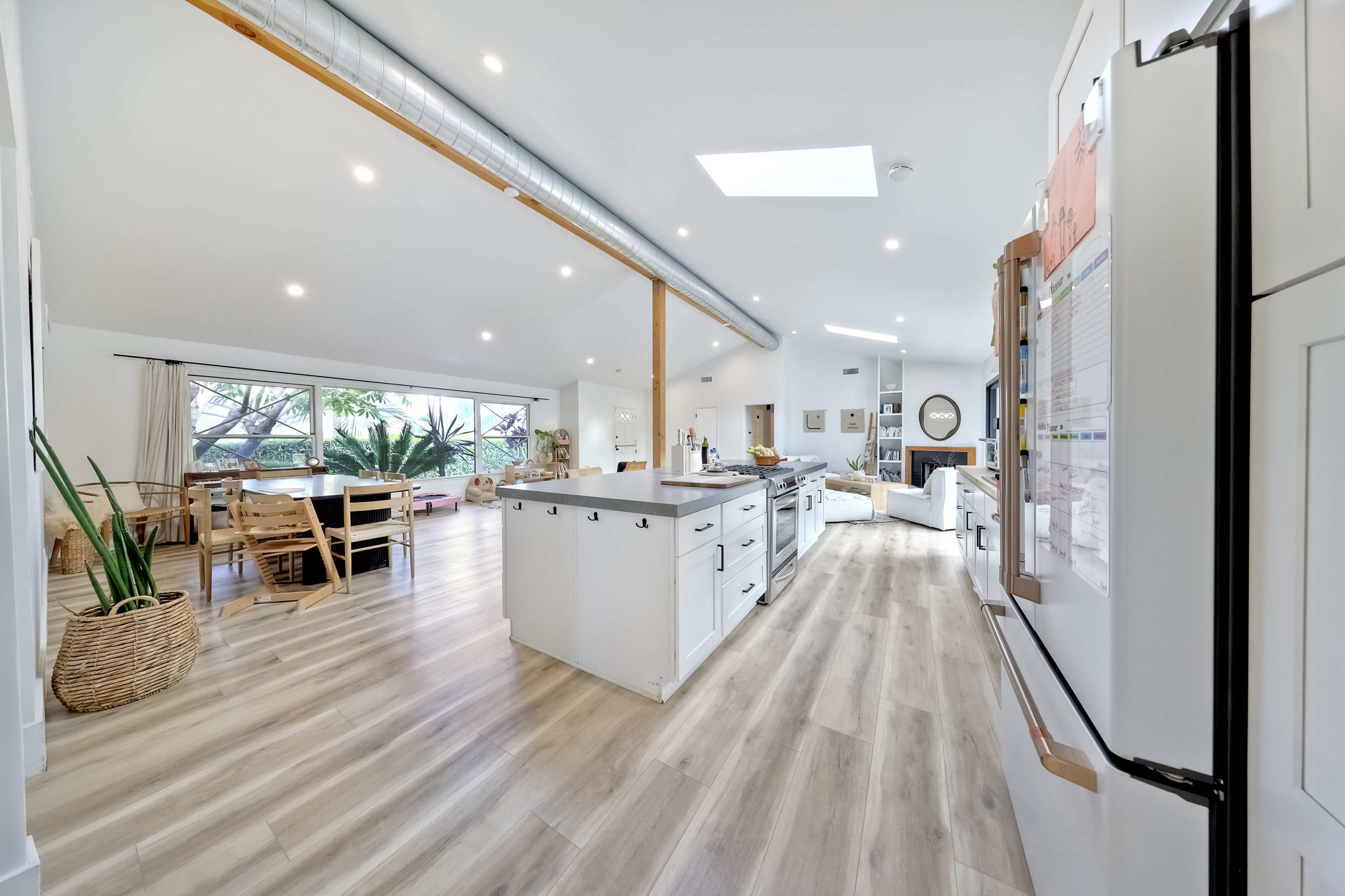The image shows a spacious, modern kitchen and dining area with wooden flooring and large windows allowing natural light to fill the room.