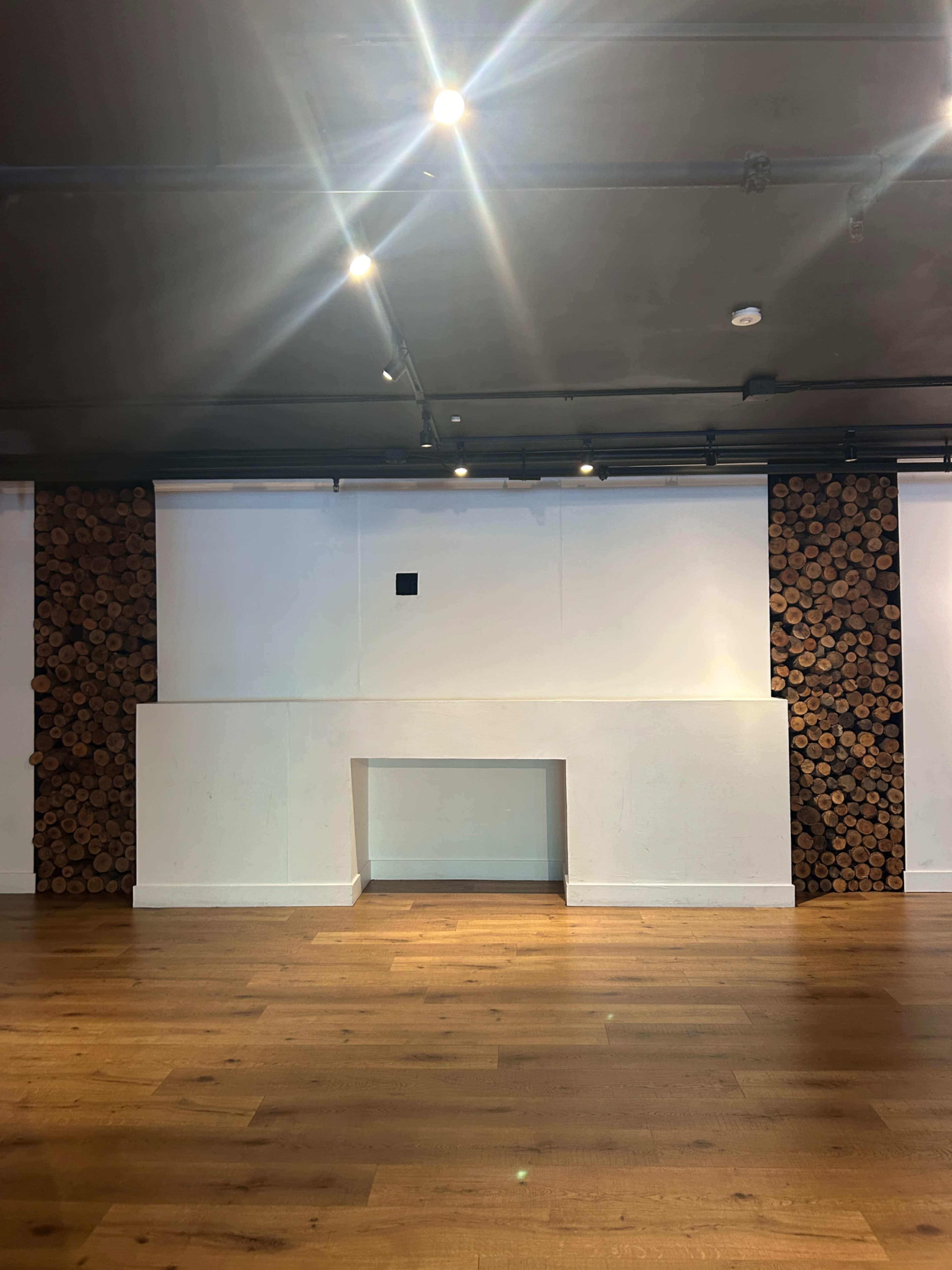 The image shows a spacious room with a wooden floor, a white wall featuring a small square inset, and two sections of stacked logs on either side.