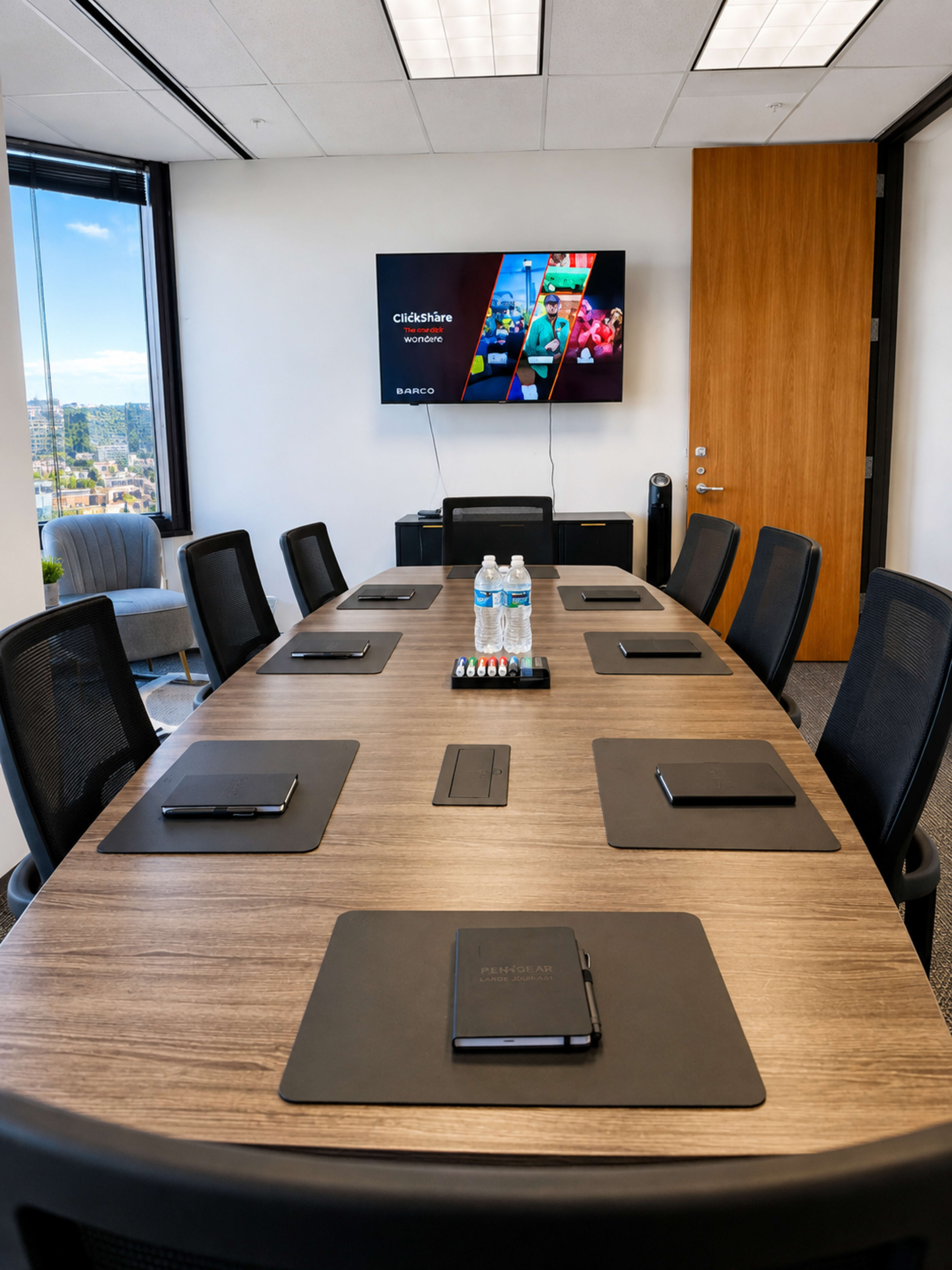 A modern conference room features a large wooden table set with notepads and pens, and a wall-mounted screen displaying a presentation titled "ClickShare."