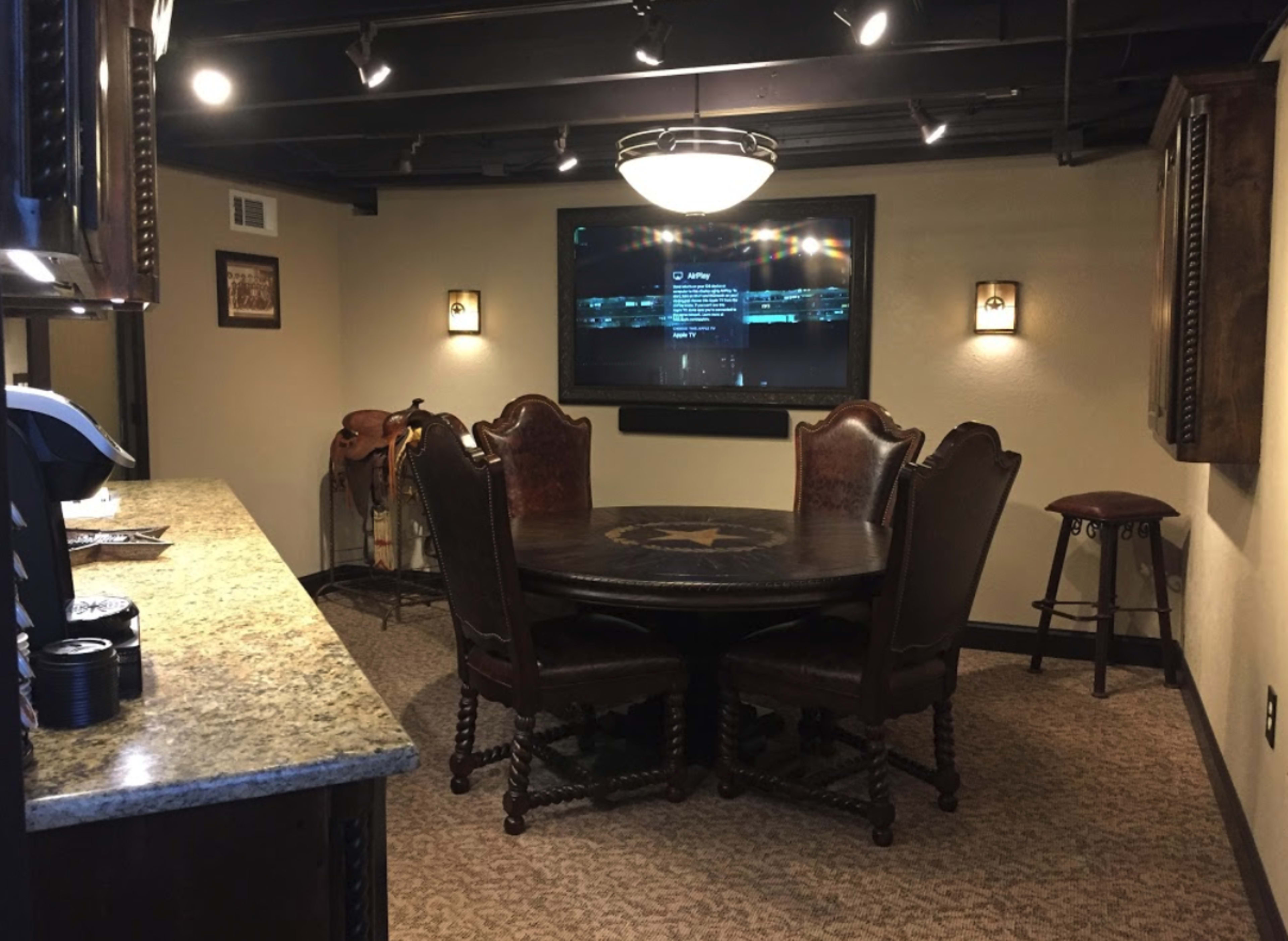 A well-lit room featuring a round table surrounded by six chairs, a mounted television on the wall, and a countertop on one side.
