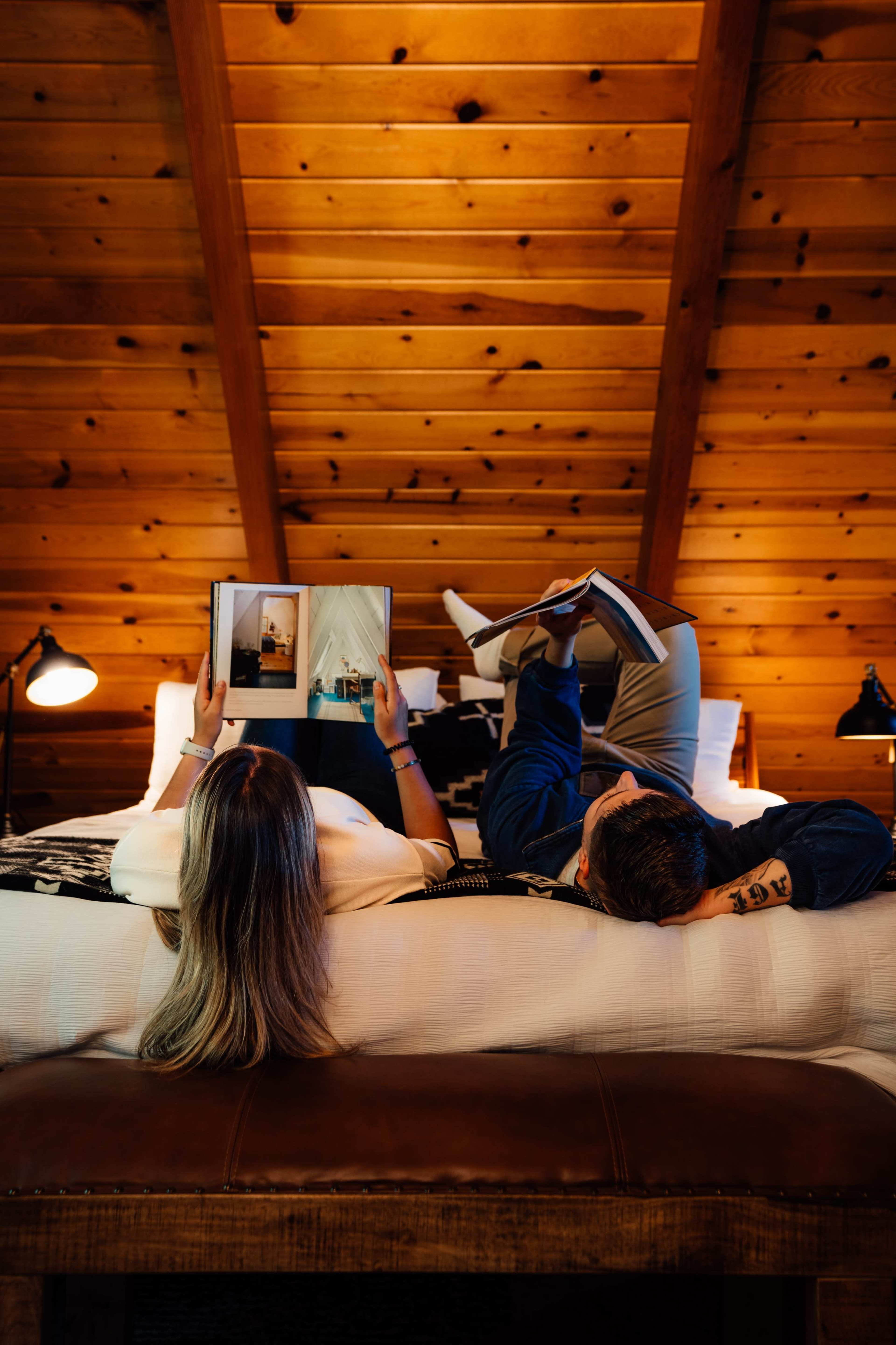 A couple lies on a bed in a wooden cabin, each reading a magazine.