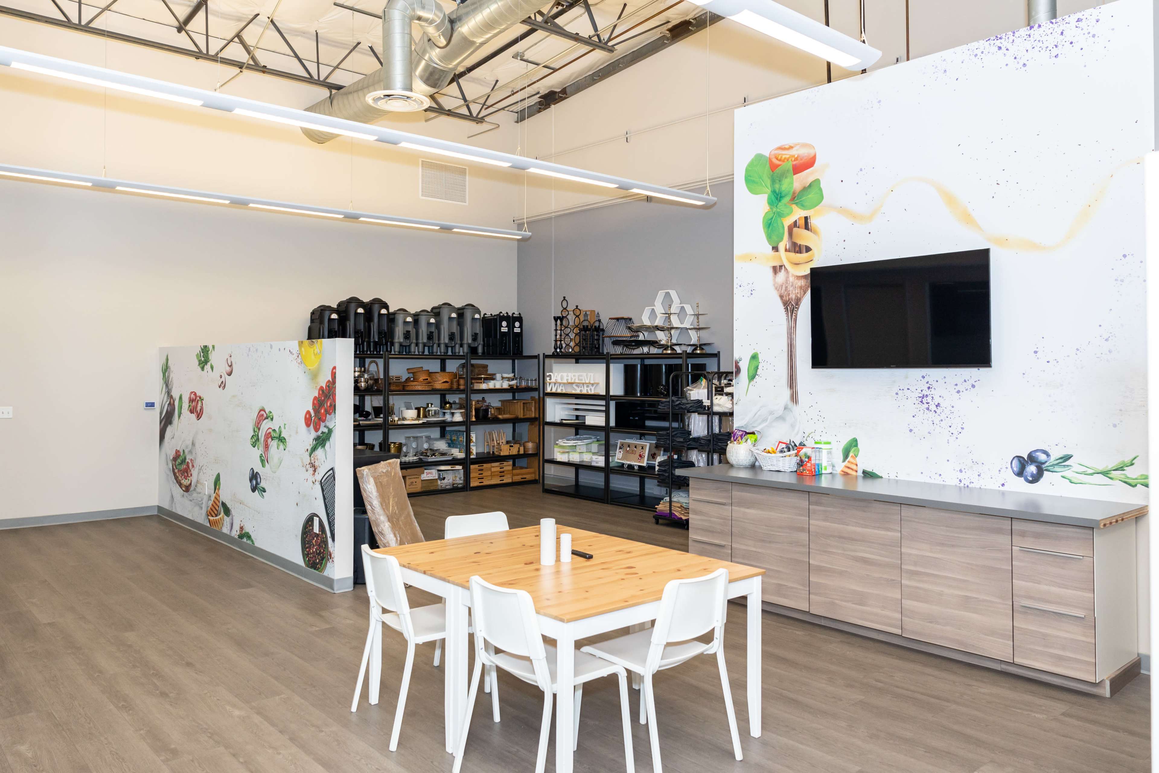 A modern kitchen space with a wooden table and white chairs, featuring various kitchen appliances and utensils organized on shelves against colorful mural walls.