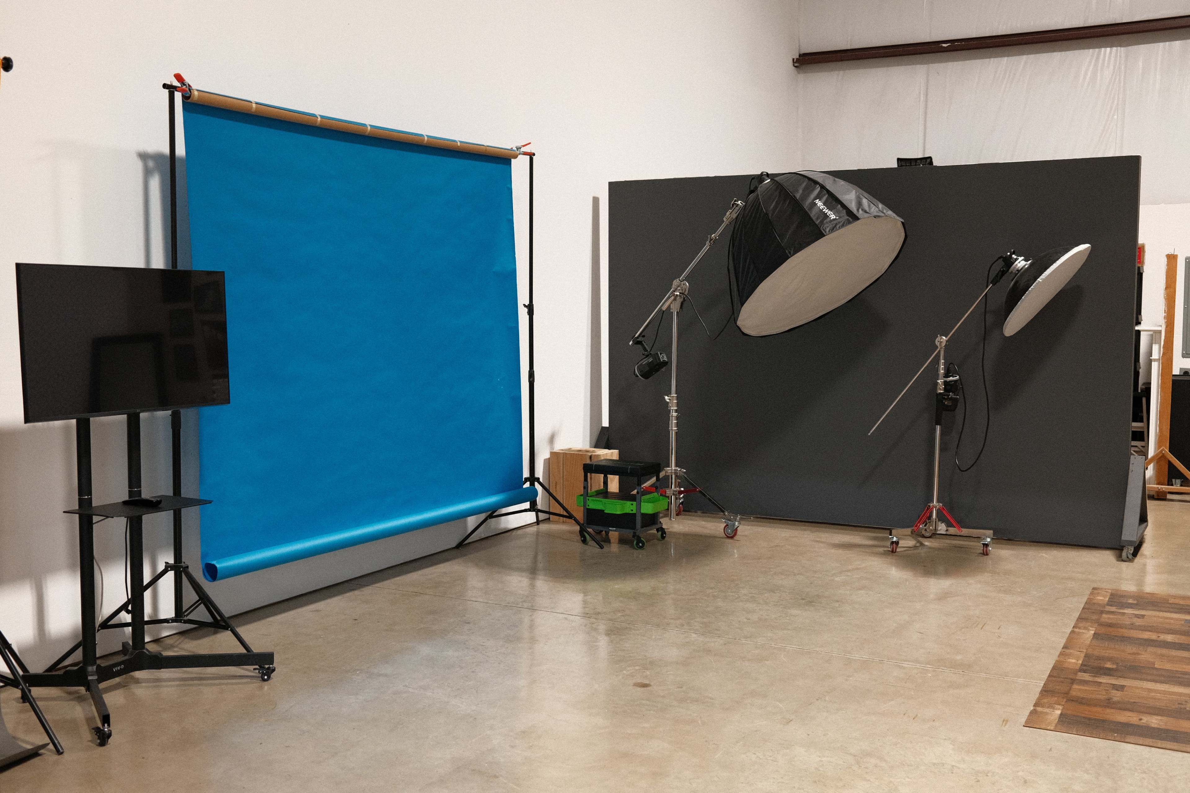 A photography studio setup featuring a blue backdrop, a large black umbrella light, and a monitor on a stand.