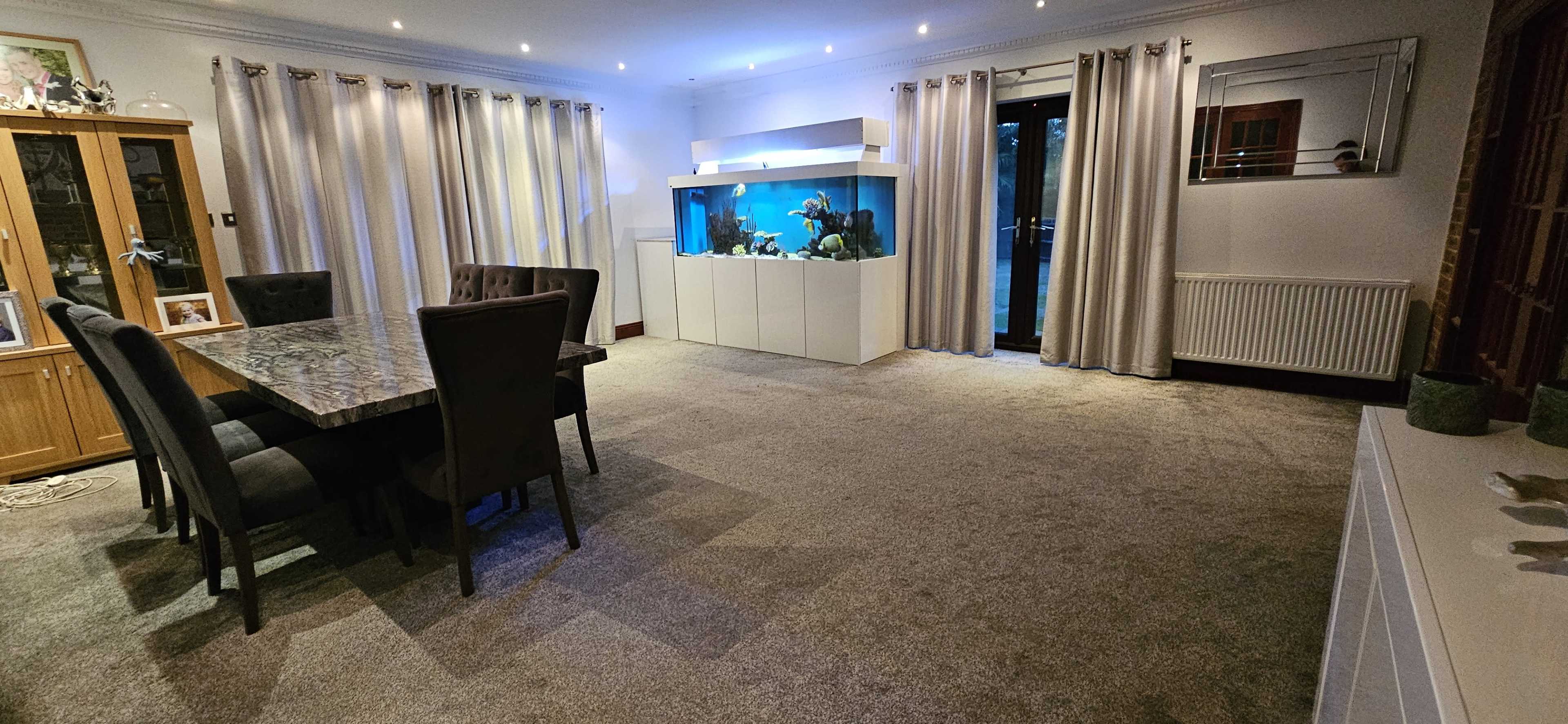 A spacious room features a dining table with six chairs, an aquarium against one wall, and curtains covering large windows.