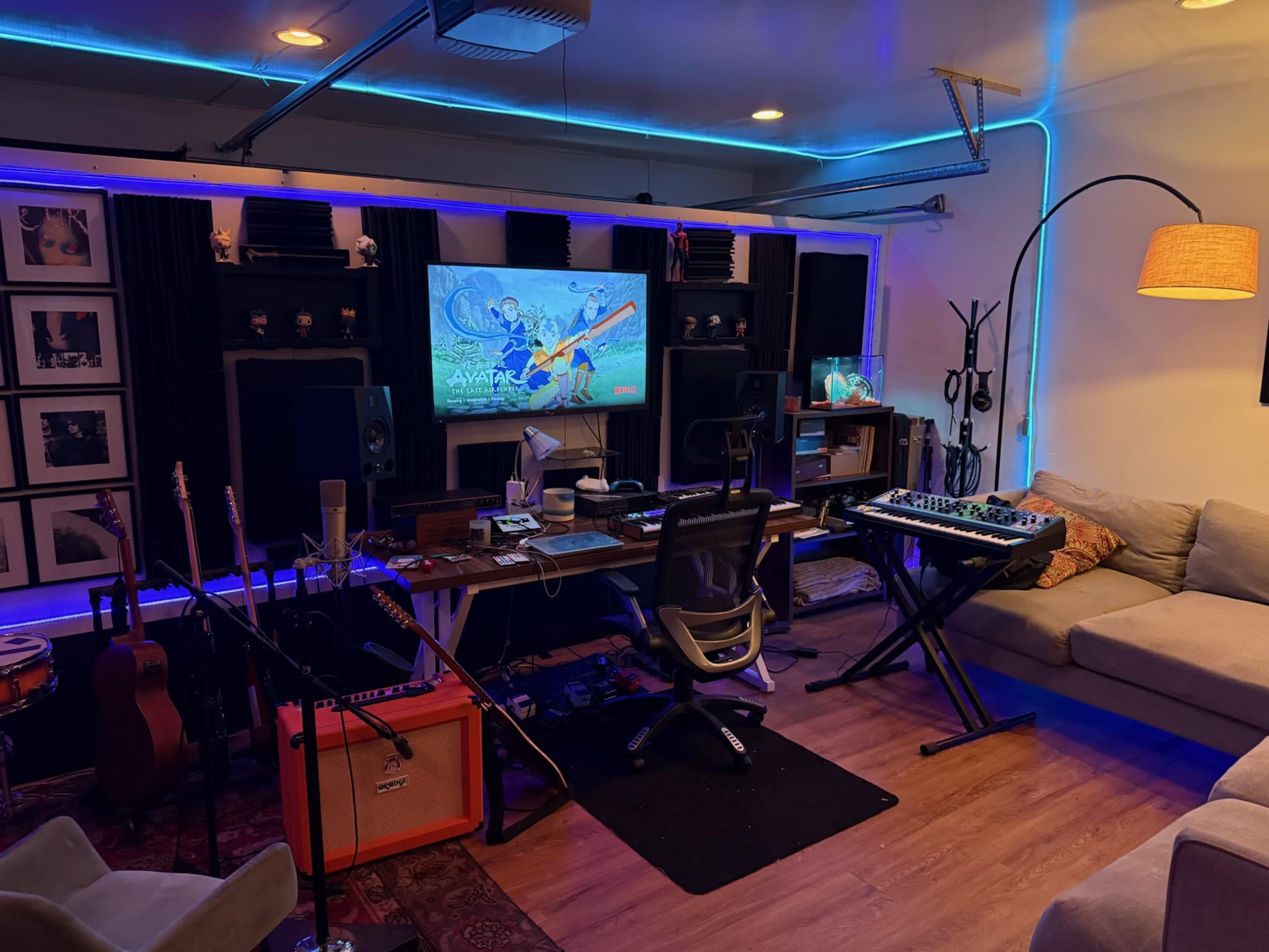 The image shows a music studio featuring a desk with a computer and audio equipment, surrounded by guitars, monitors, and illuminated by blue LED lights.