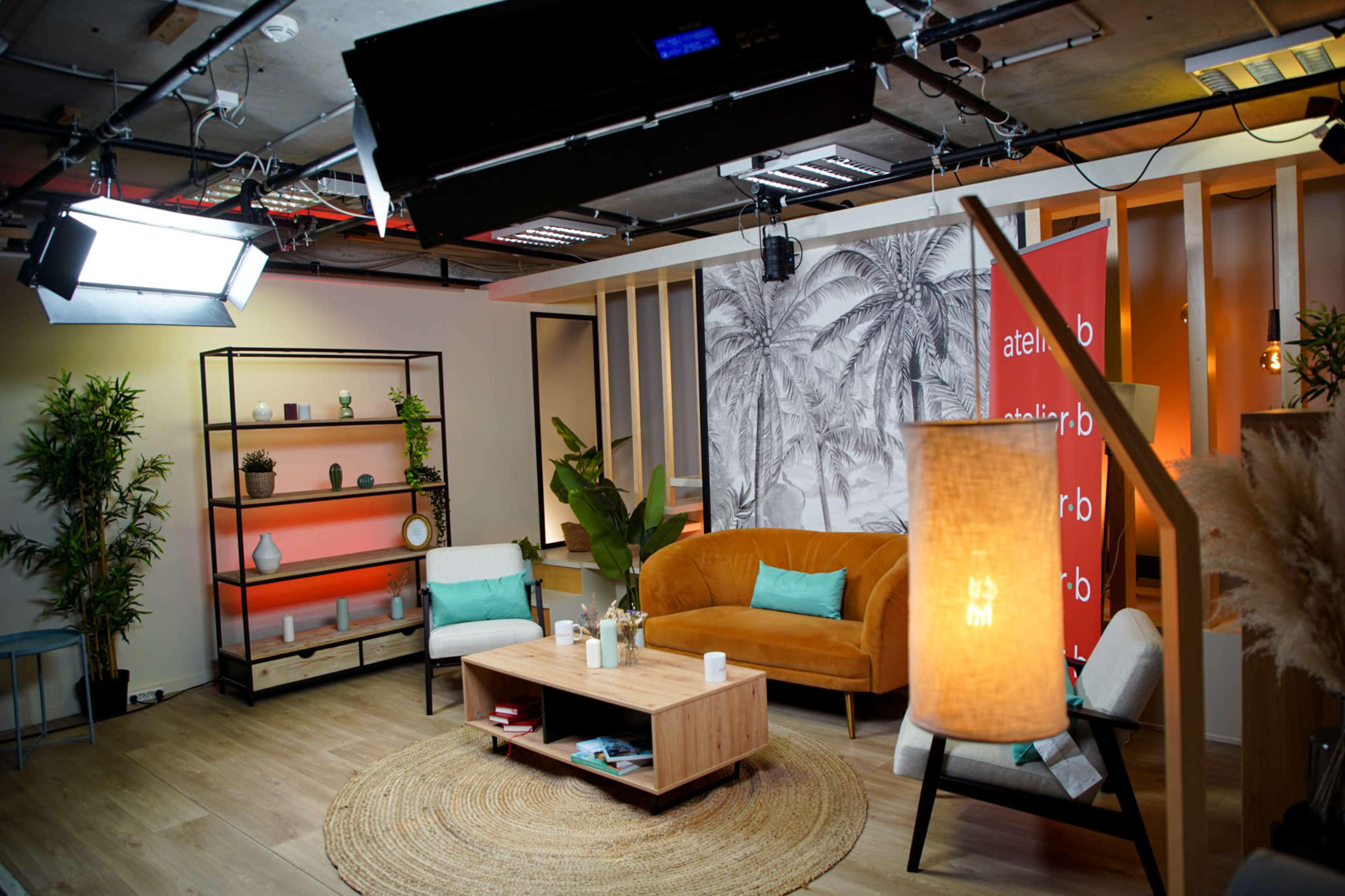The image shows a modern studio setting with a cozy seating area featuring an orange sofa, two armchairs, a coffee table, and decorative plants, all under studio lighting.
