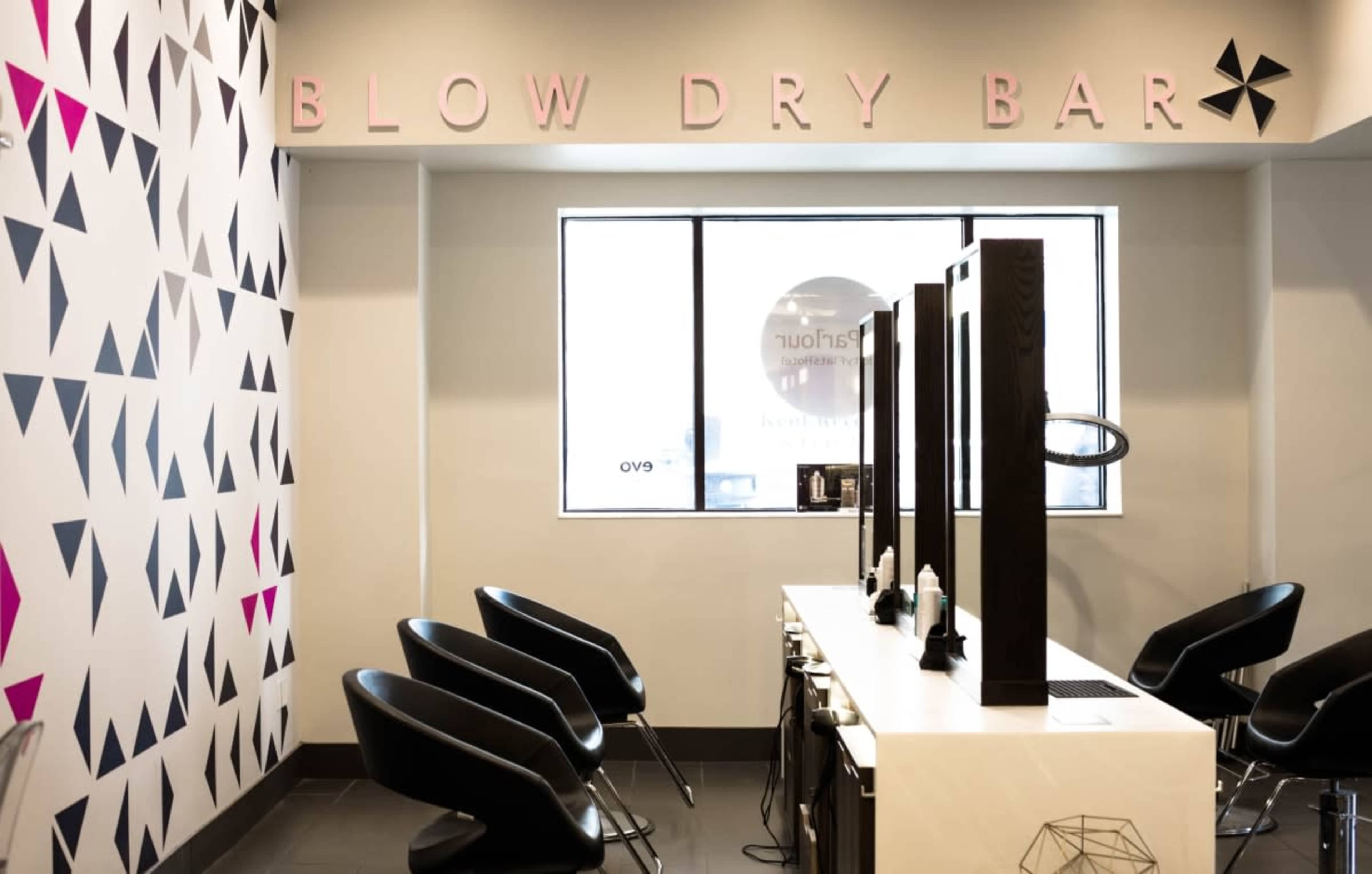 The interior of a blow dry bar featuring a decorative wall, black salon chairs, and a row of stylist stations with mirrors.