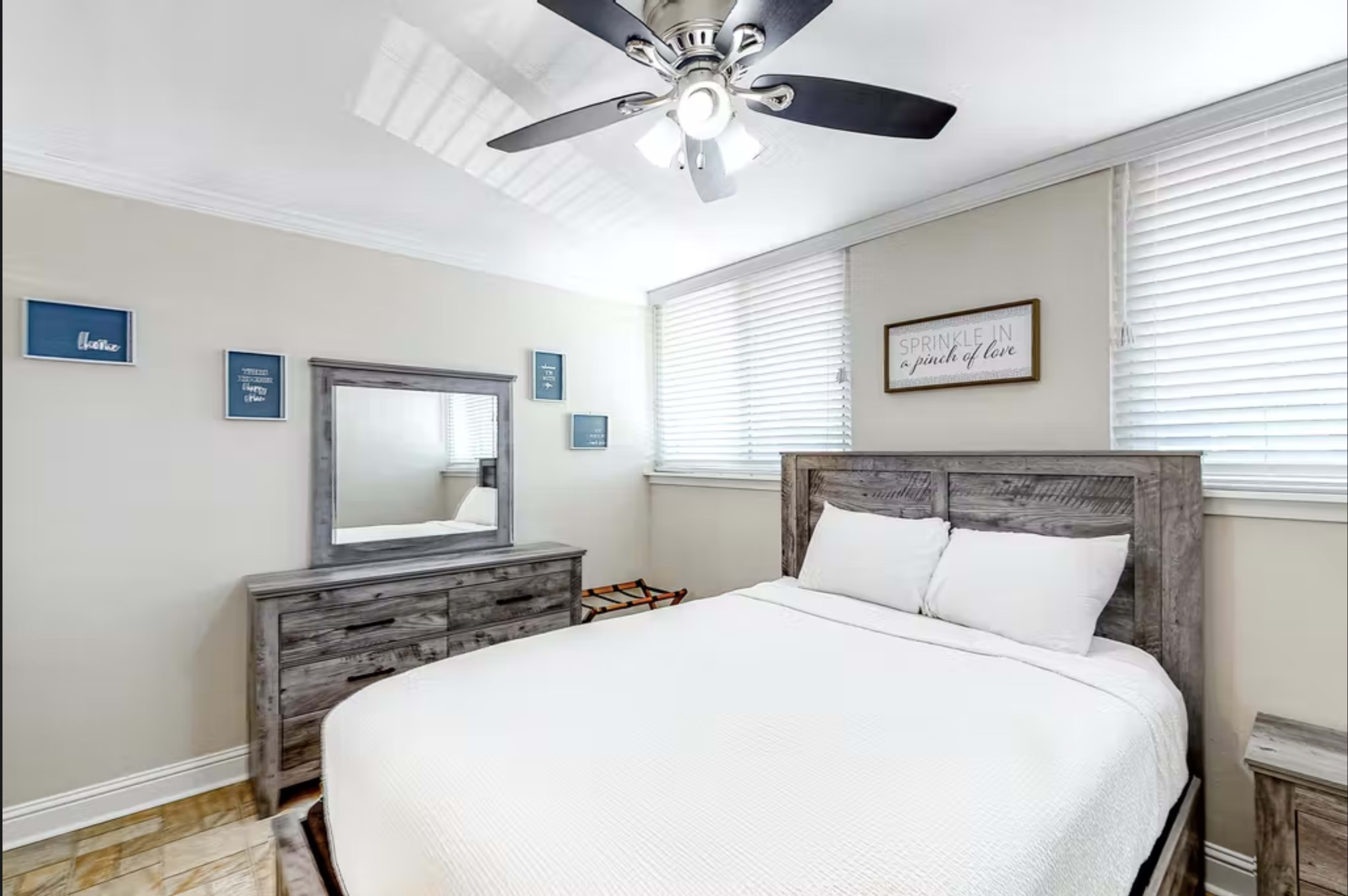 A well-lit bedroom featuring a double bed, a wooden dresser, a mirror, and several wall decorations.