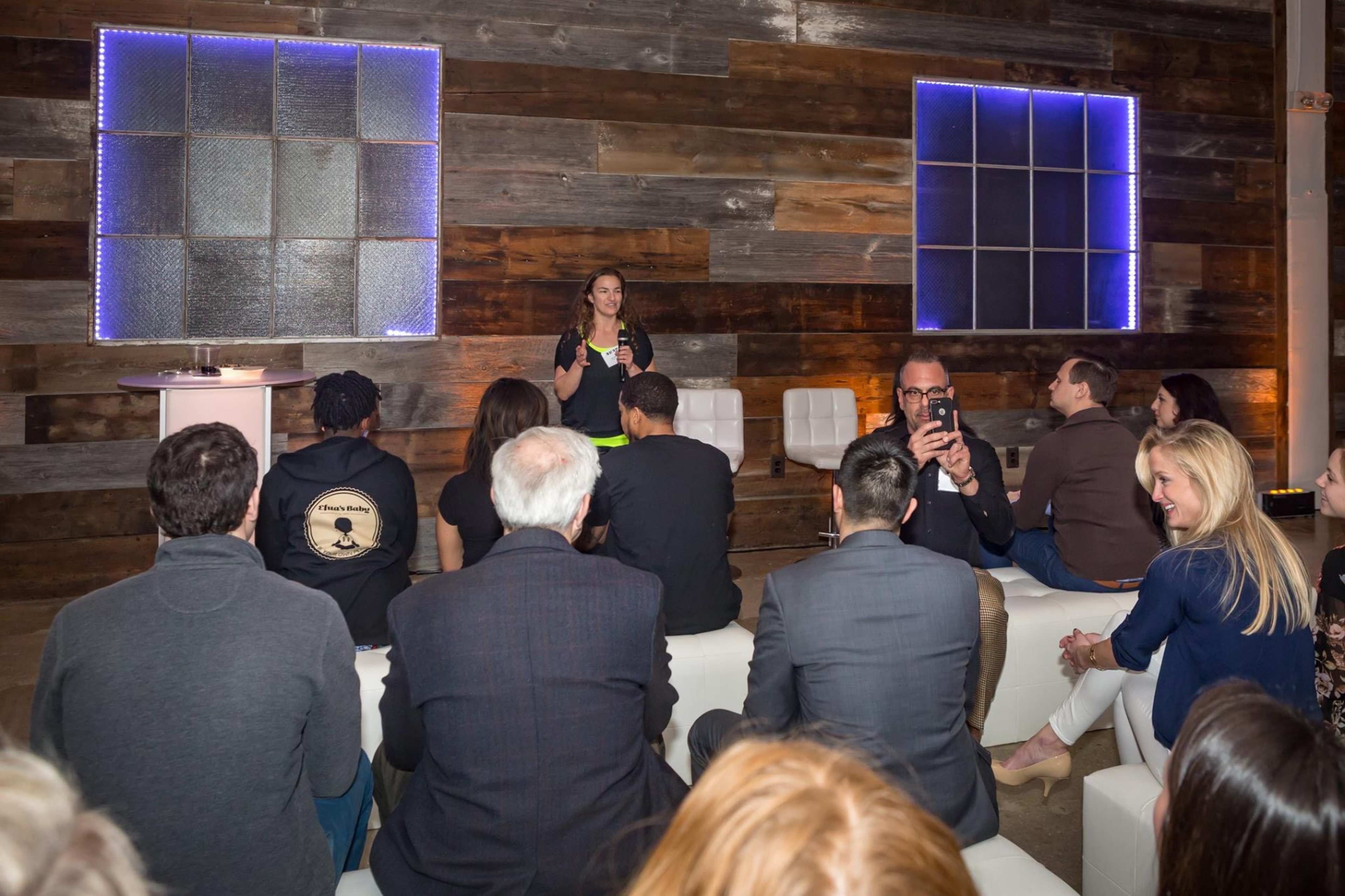 A speaker addresses an audience seated on white sofas in a stylish interior with wooden walls and glowing blue lights.