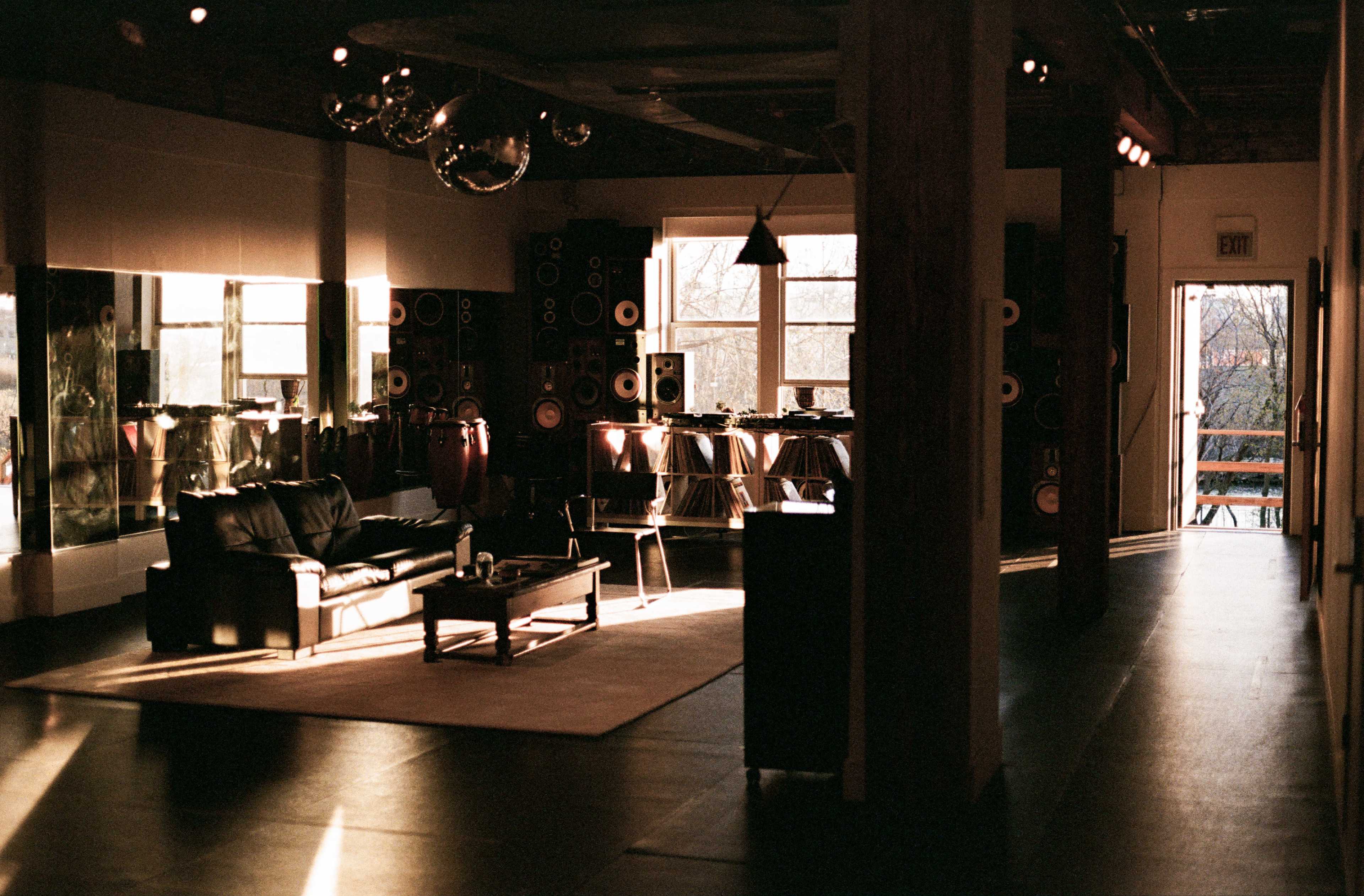 The image shows a spacious room featuring a black sofa, a wooden coffee table, and large speakers lining the walls, with sunlight streaming through windows and illuminating the space.