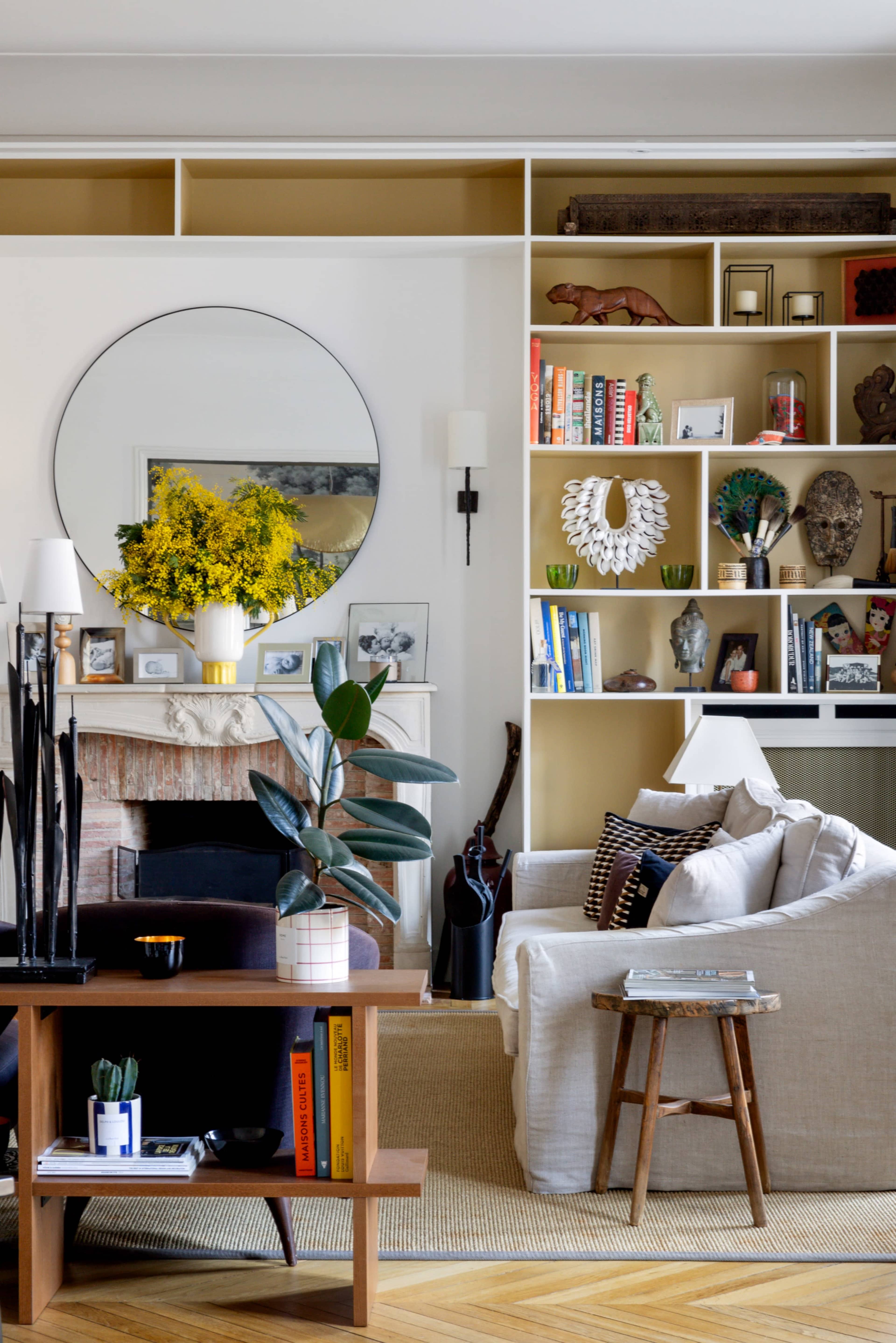 The image shows a bright living room featuring a light-colored sofa, a round mirror above a mantel, and shelves filled with various decorative items and books.