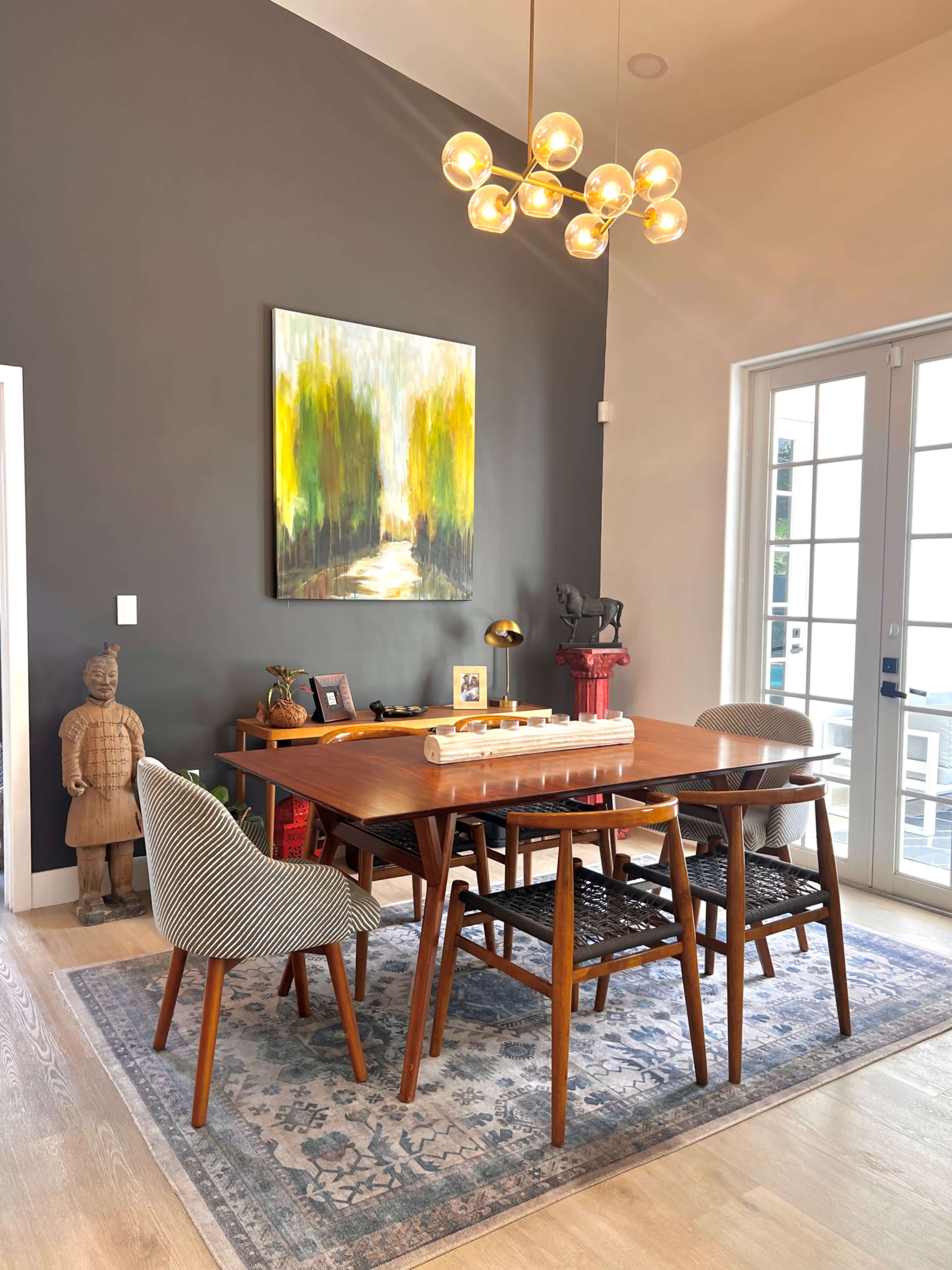 A modern dining area features a wooden table surrounded by chairs, a large painting on the wall, and decorative items including a sculpture and a vintage piece.