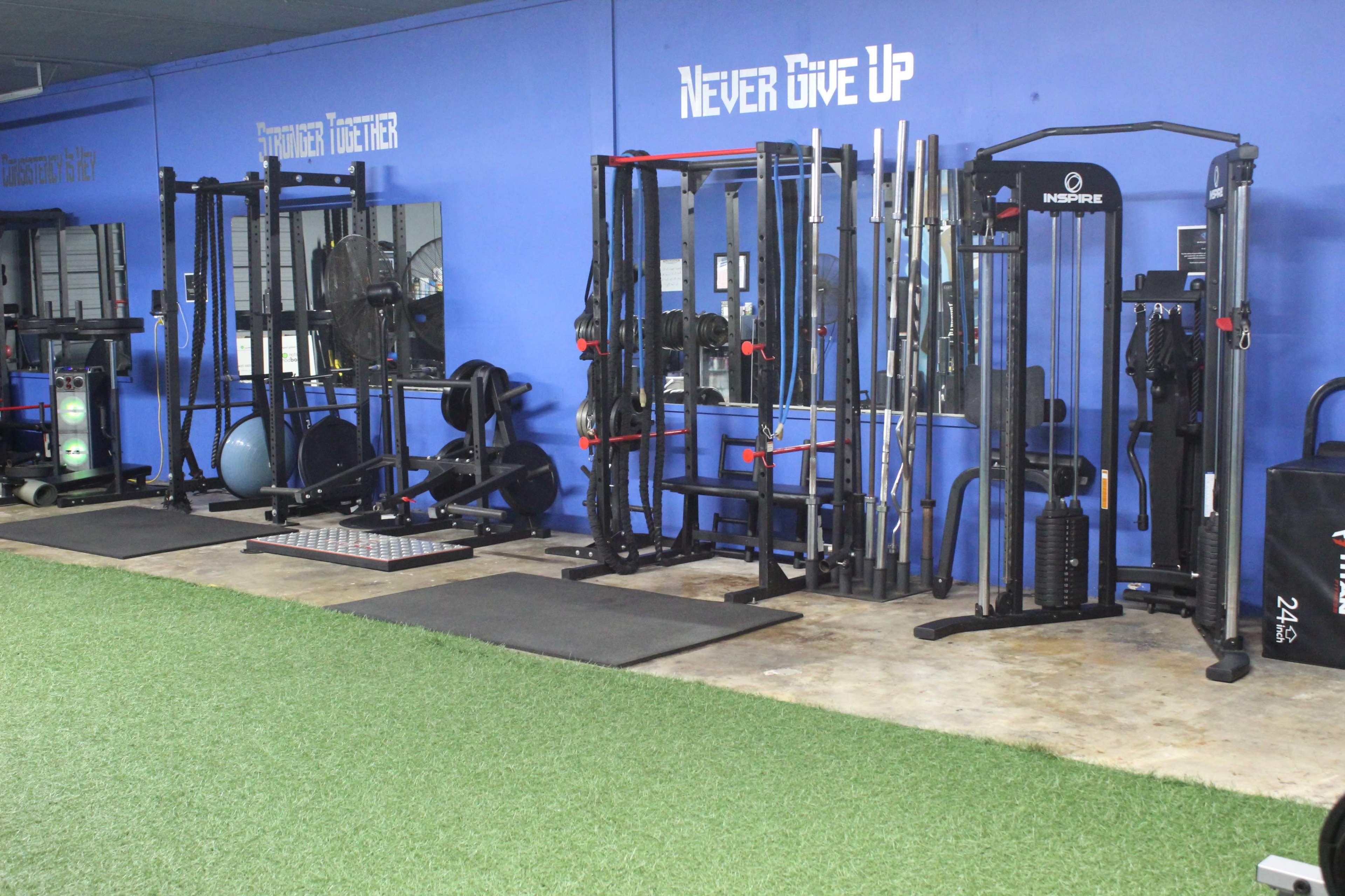 A gym interior with various exercise equipment and weights arranged along a blue wall, featuring motivational text "Never Give Up."