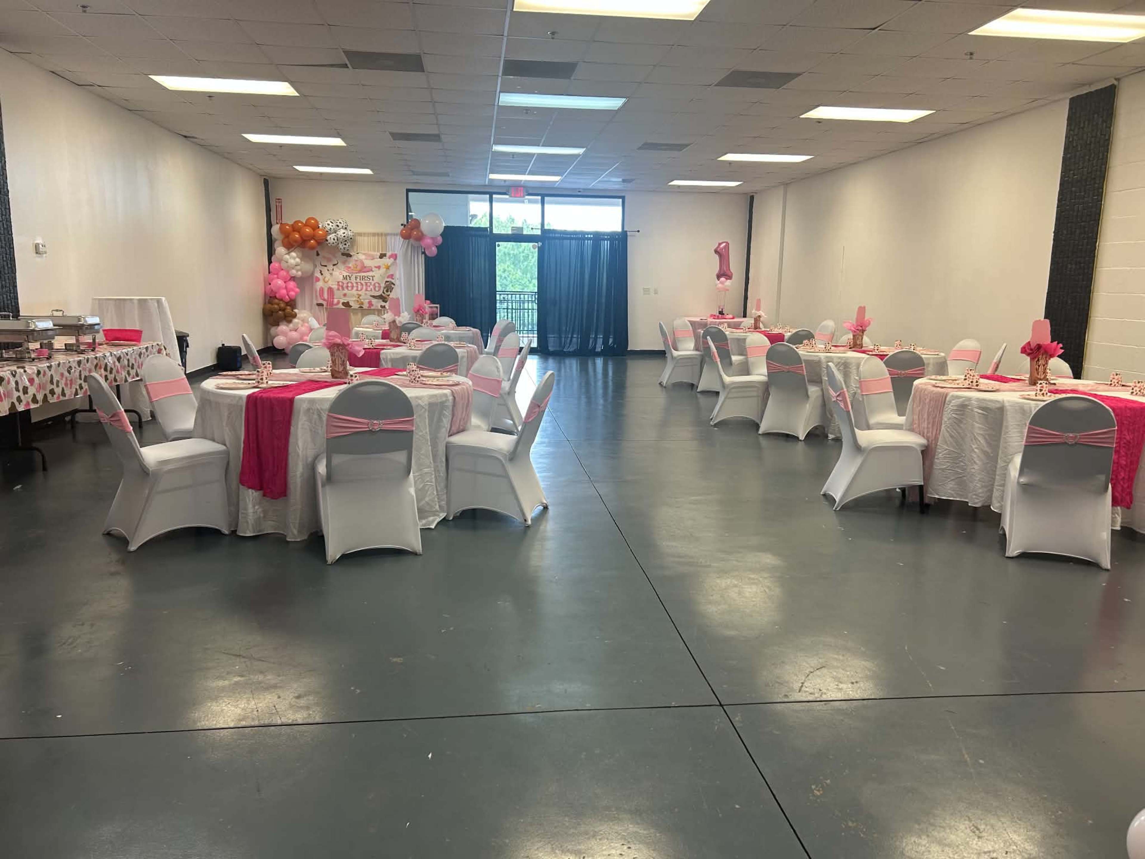 The image shows a decorated event space with multiple round tables set for a celebration, featuring pink and white table settings and balloons in the background.