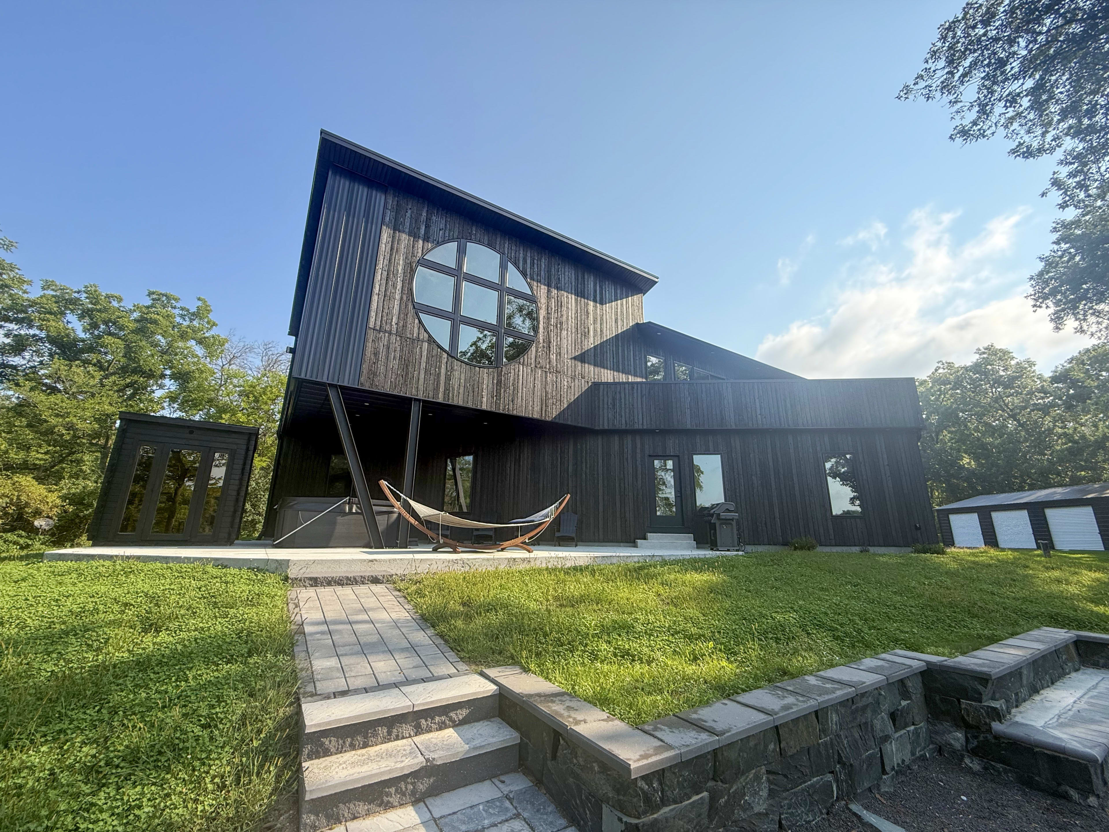 The image shows a modern, black wooden house with a large circular window, a stone pathway, and a hammock in the front yard surrounded by greenery.