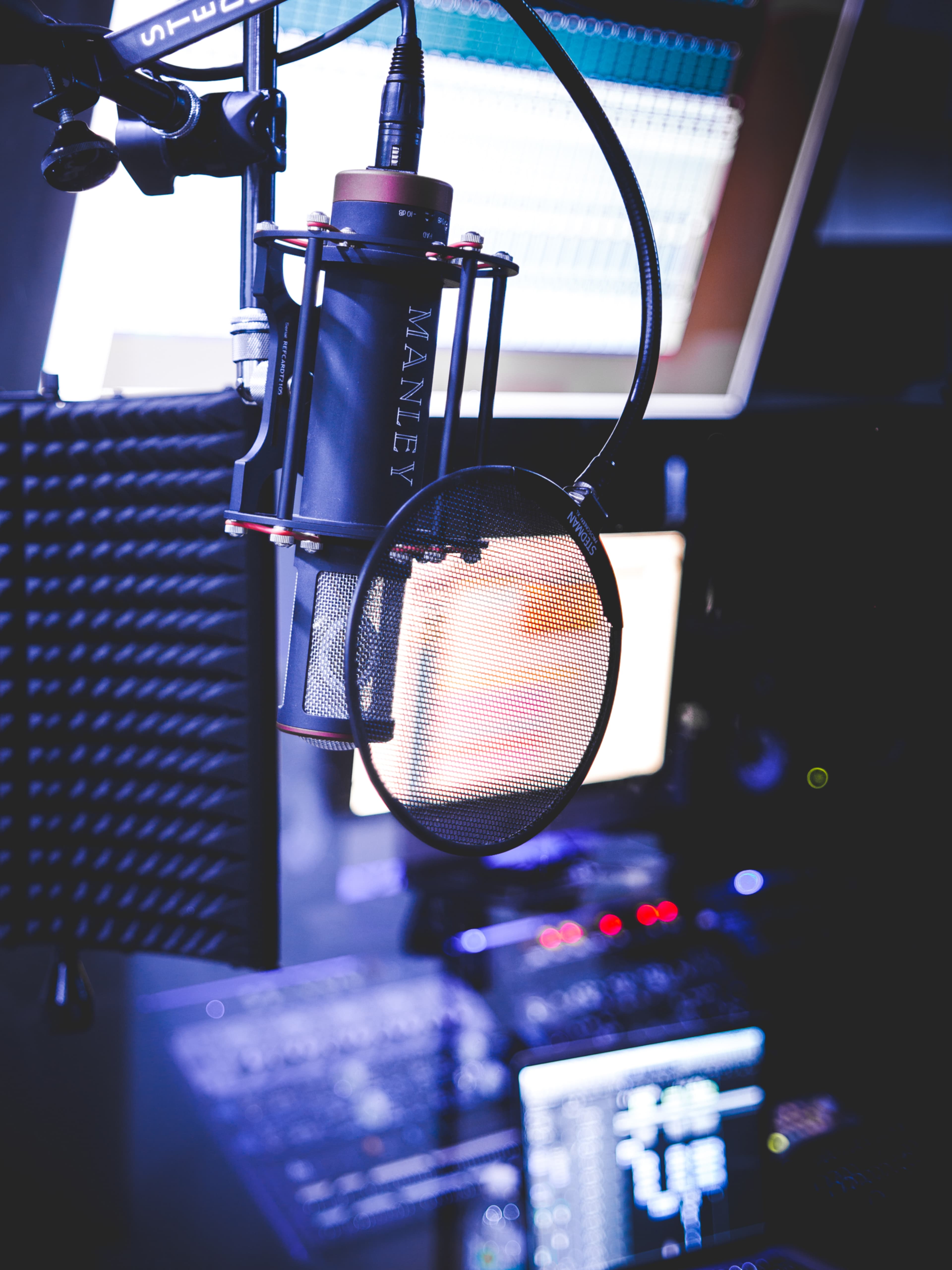 A studio microphone is positioned in front of sound equipment and computer monitors in a recording environment.