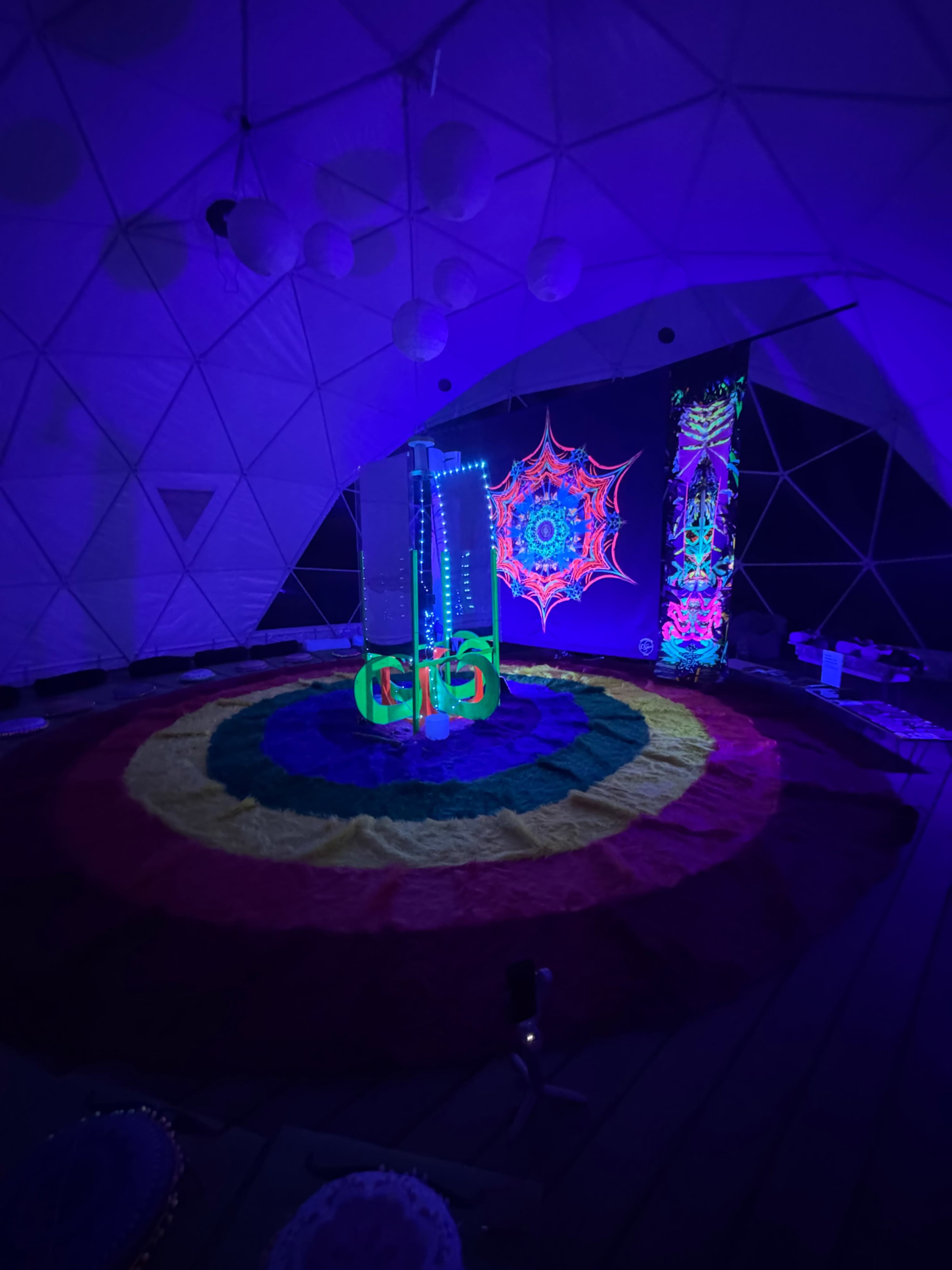 The interior of a geodesic dome is illuminated by colorful lights, featuring a vibrant circular rug at the center and decorative elements on the walls.