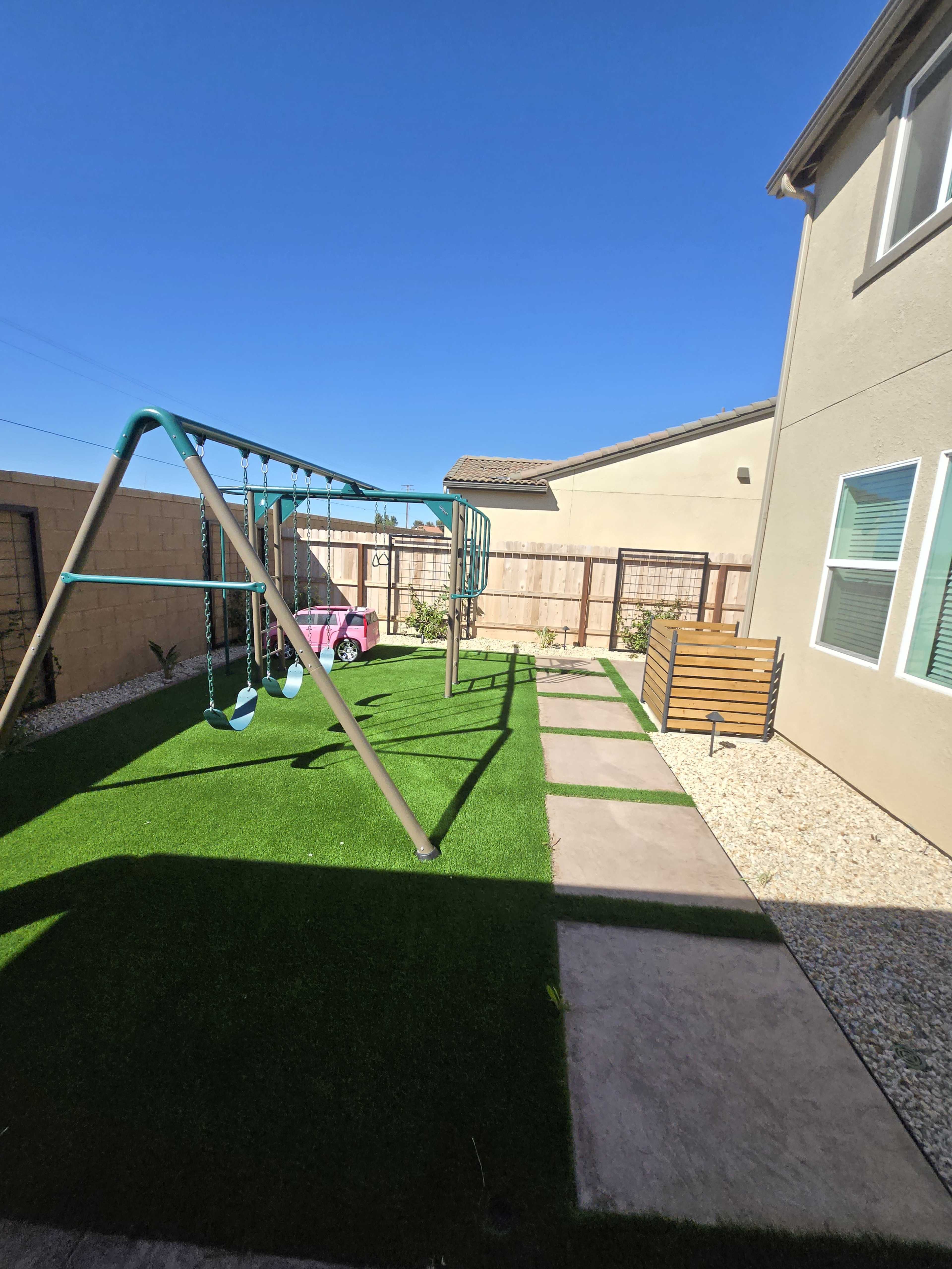 The image shows a backyard featuring a playground set with swings, a paved pathway, artificial grass, and a small wooden bench near the house.