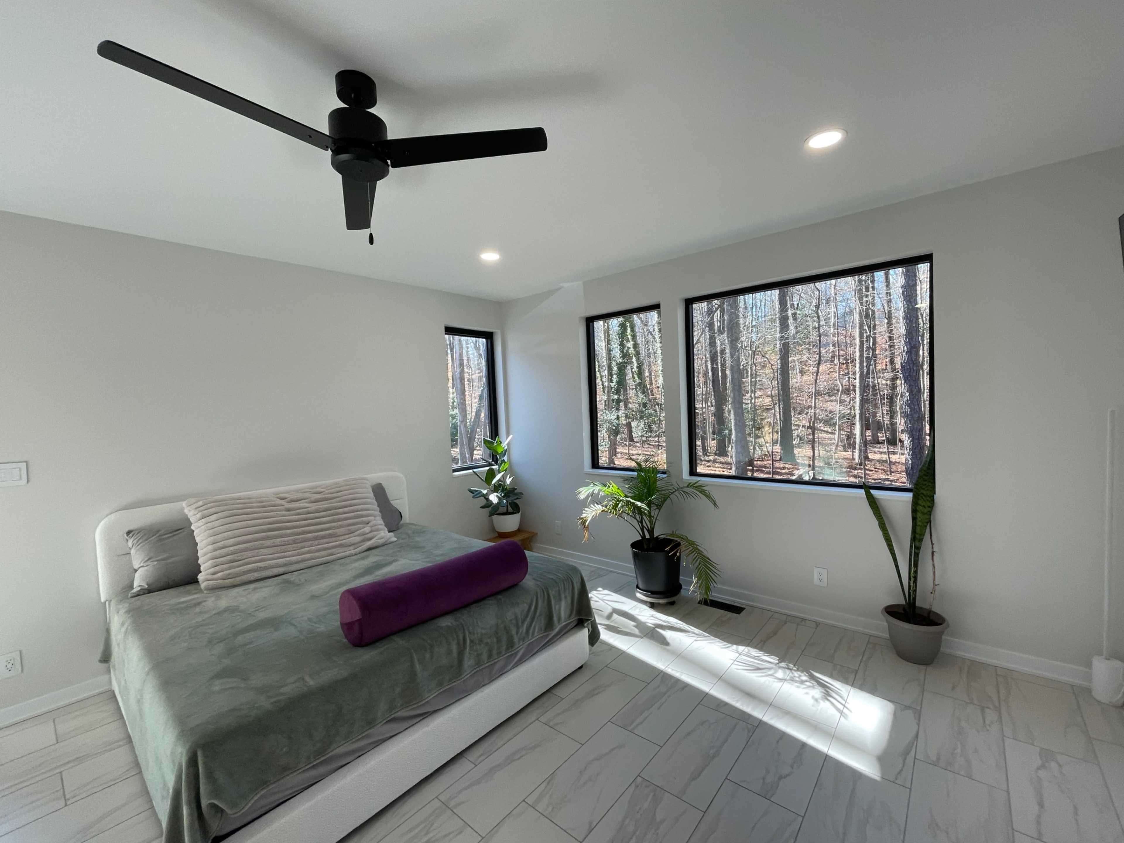 A modern bedroom features a bed with a purple bolster pillow, surrounded by large windows offering a view of the wooded area outside.