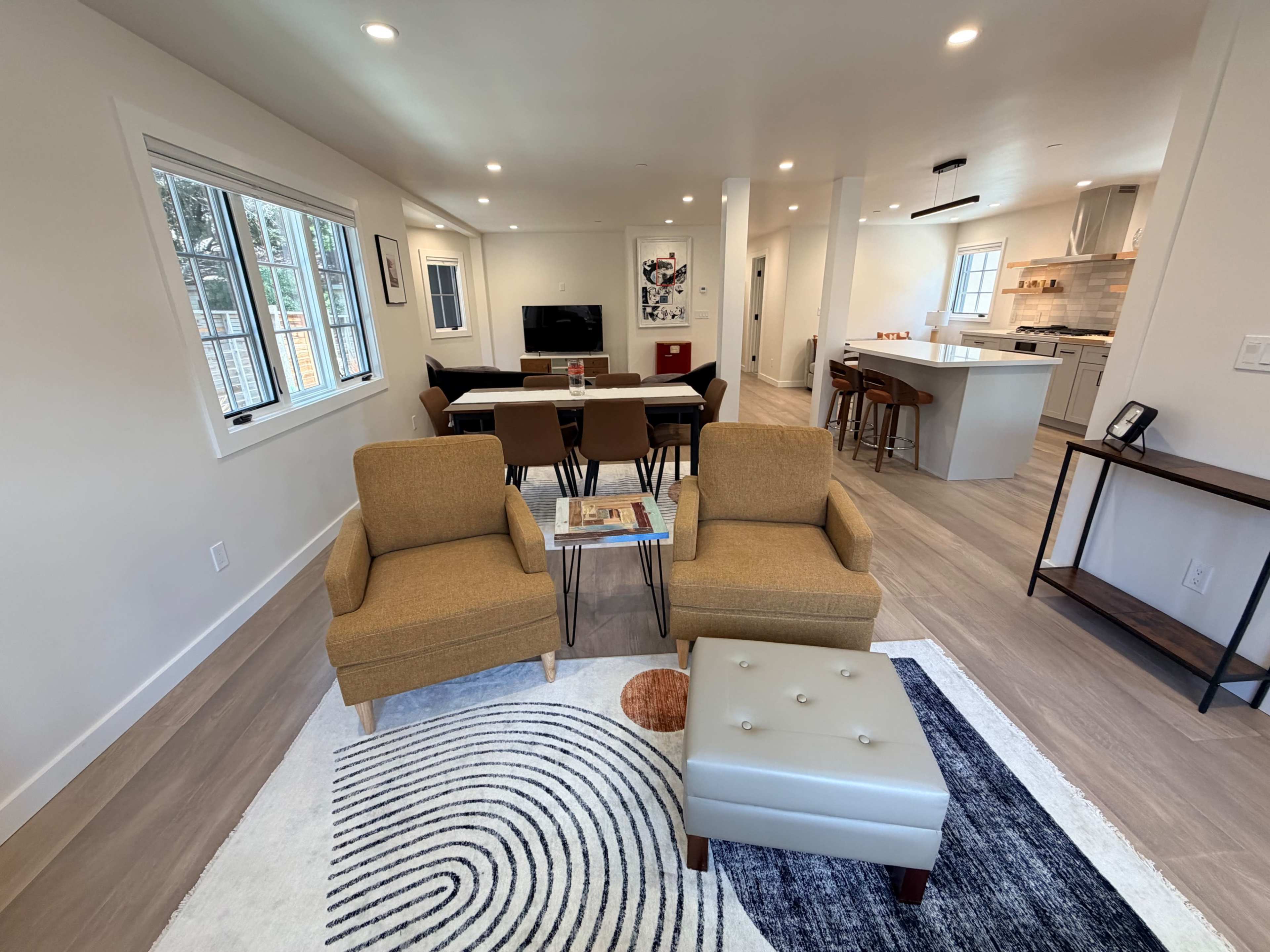 The image shows a modern living space with two tan armchairs and a small table, facing an open kitchen and dining area.