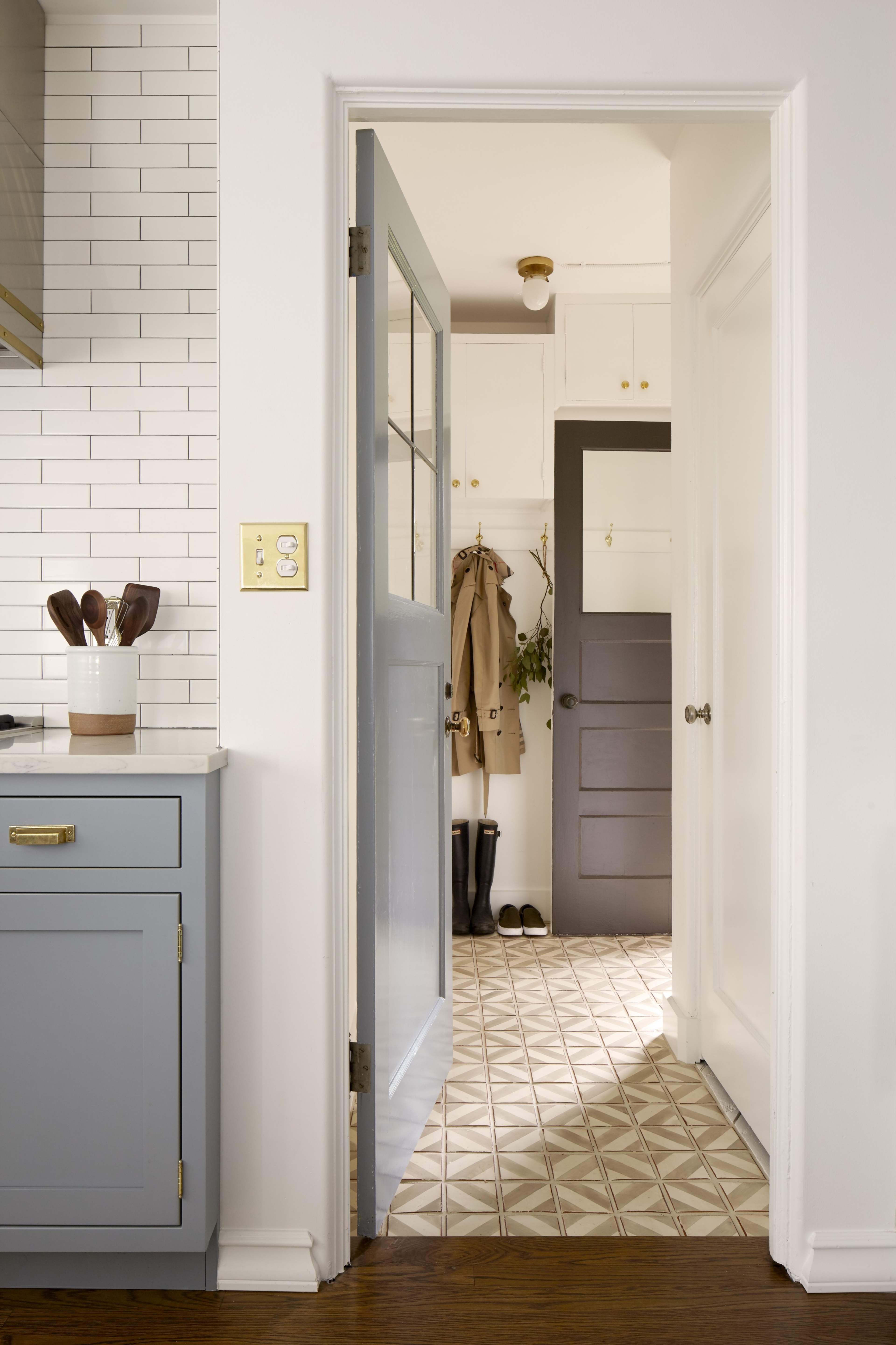 A view through a doorway reveals a kitchen with light gray cabinets and a patterned floor, leading to a hallway with a coat and boots.