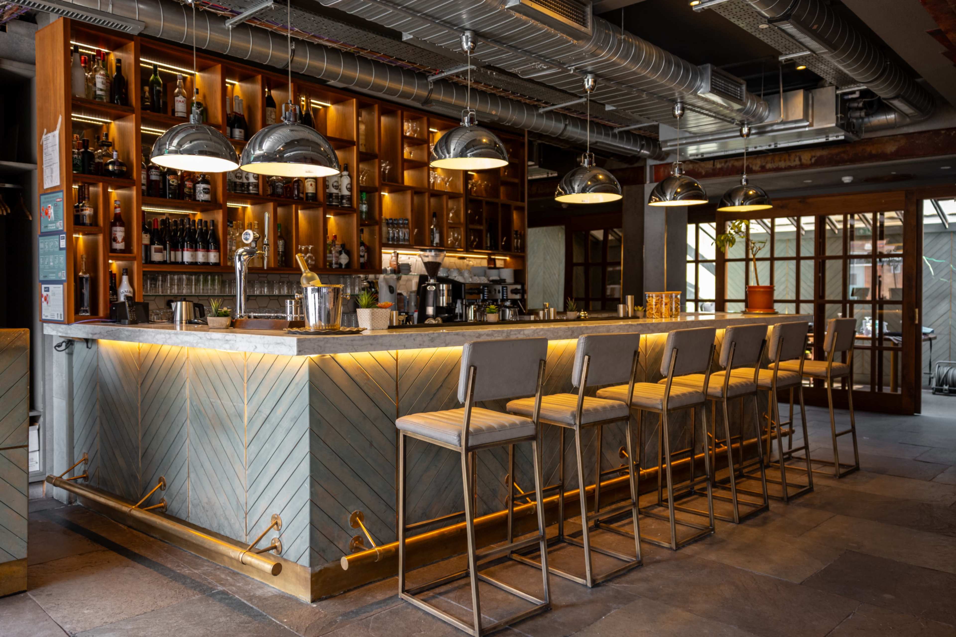 The image shows a modern bar with a well-lit counter, high stools, and shelves stocked with various bottles and glasses.