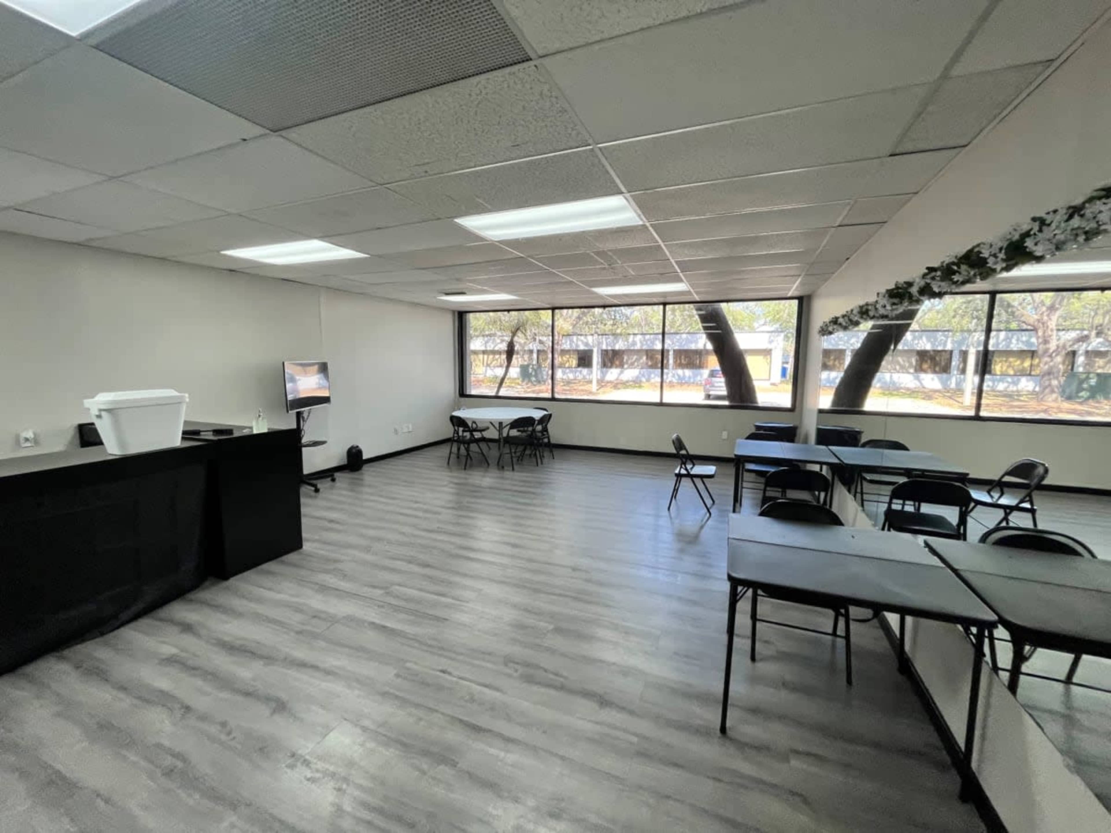 The image shows a sparsely furnished meeting room with several black chairs and tables, a white container, and large windows letting in natural light.