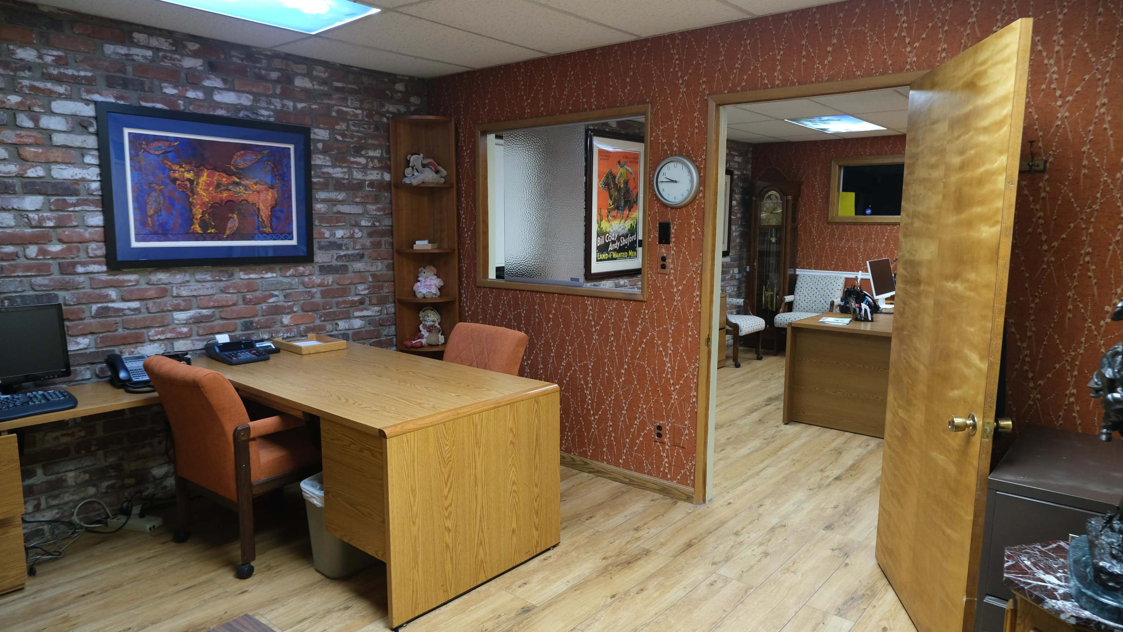 An office space with wooden desks, brick walls, and a visible adjacent room containing a clock and artwork.