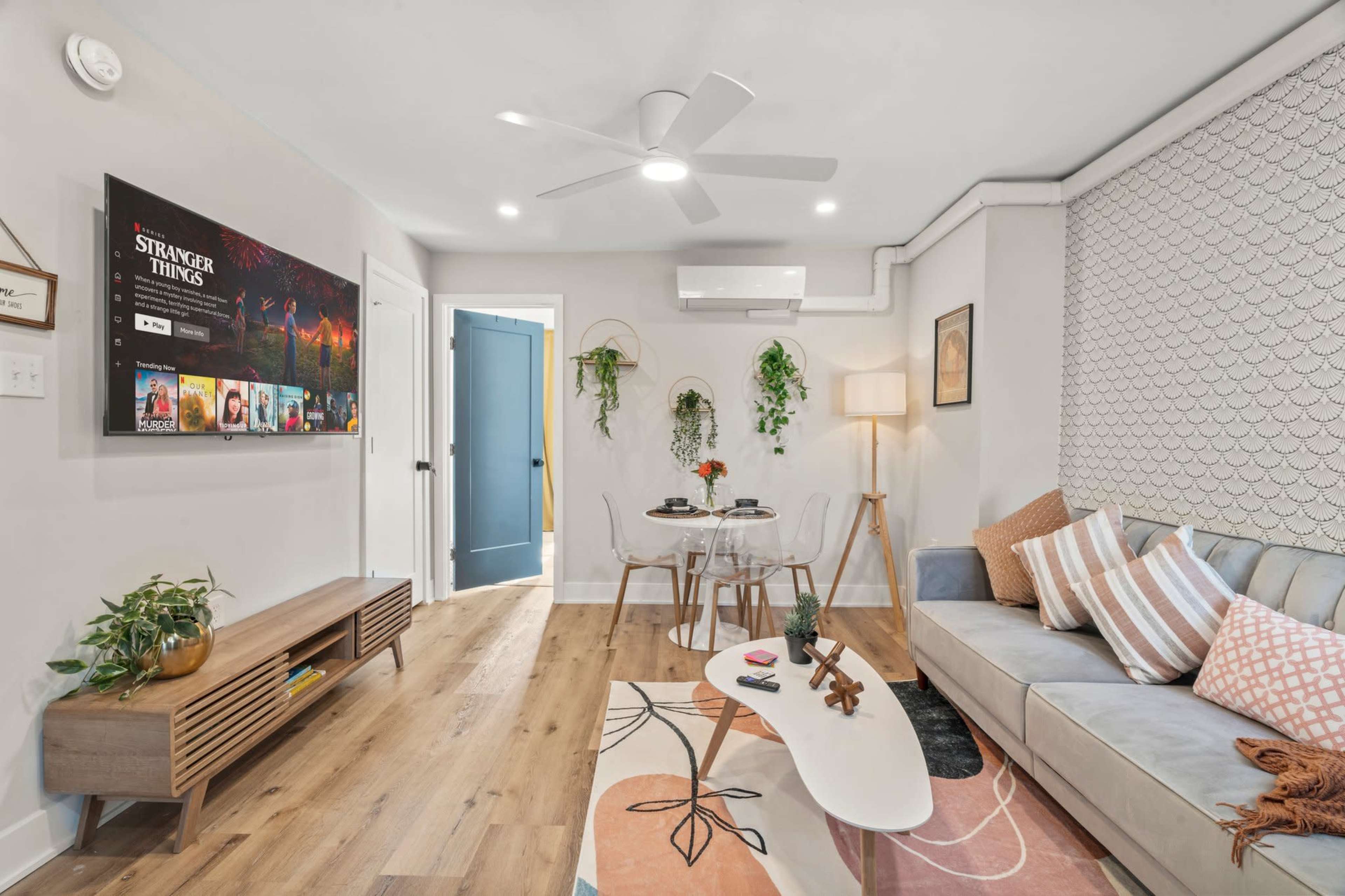 The image shows a modern living room featuring a light-colored sofa, a wooden media console with a large television displaying a series, and a decorative coffee table.