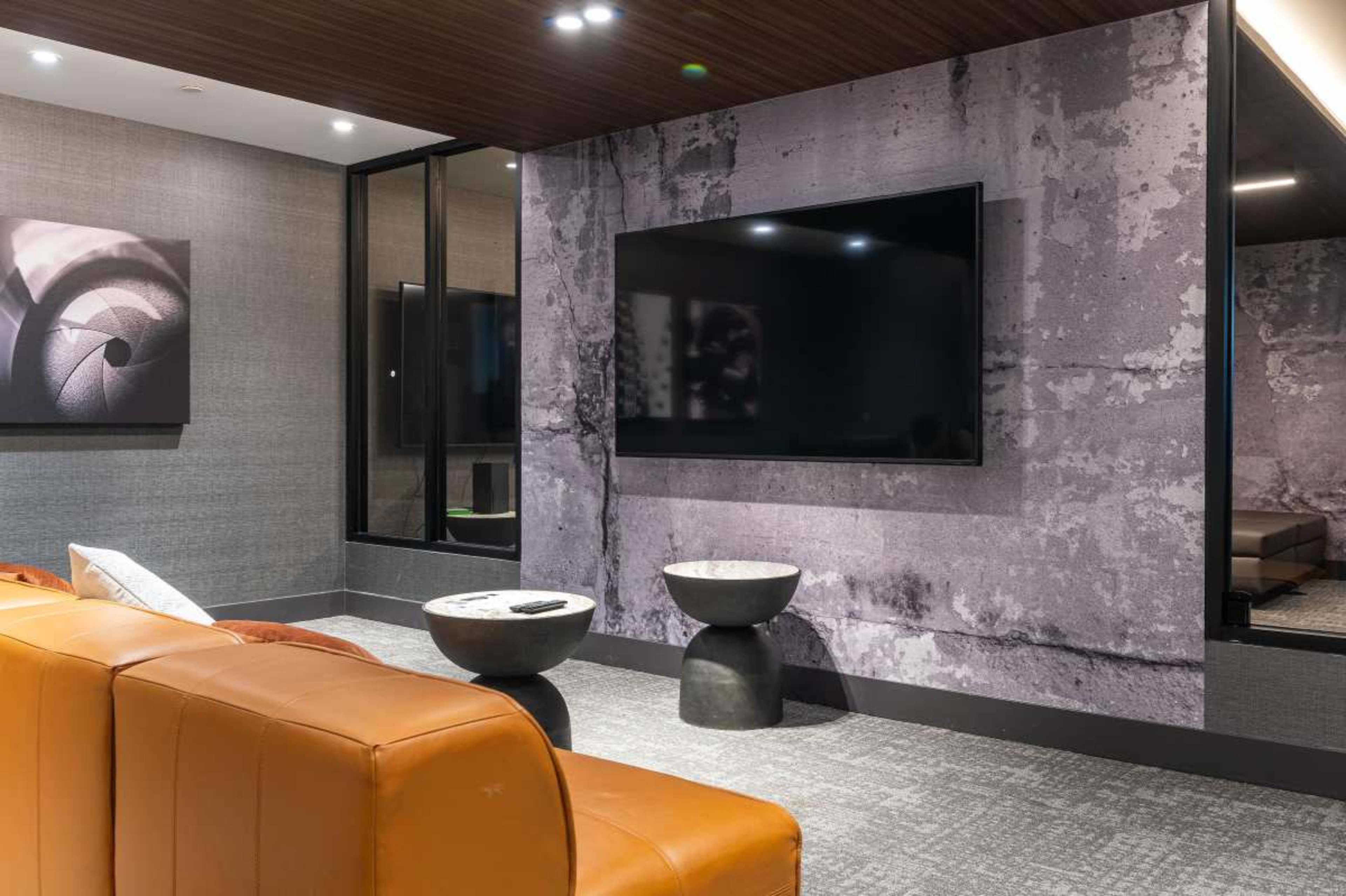 The image shows a modern lounge area featuring a large television mounted on a textured wall, with a brown leather couch and round side tables.
