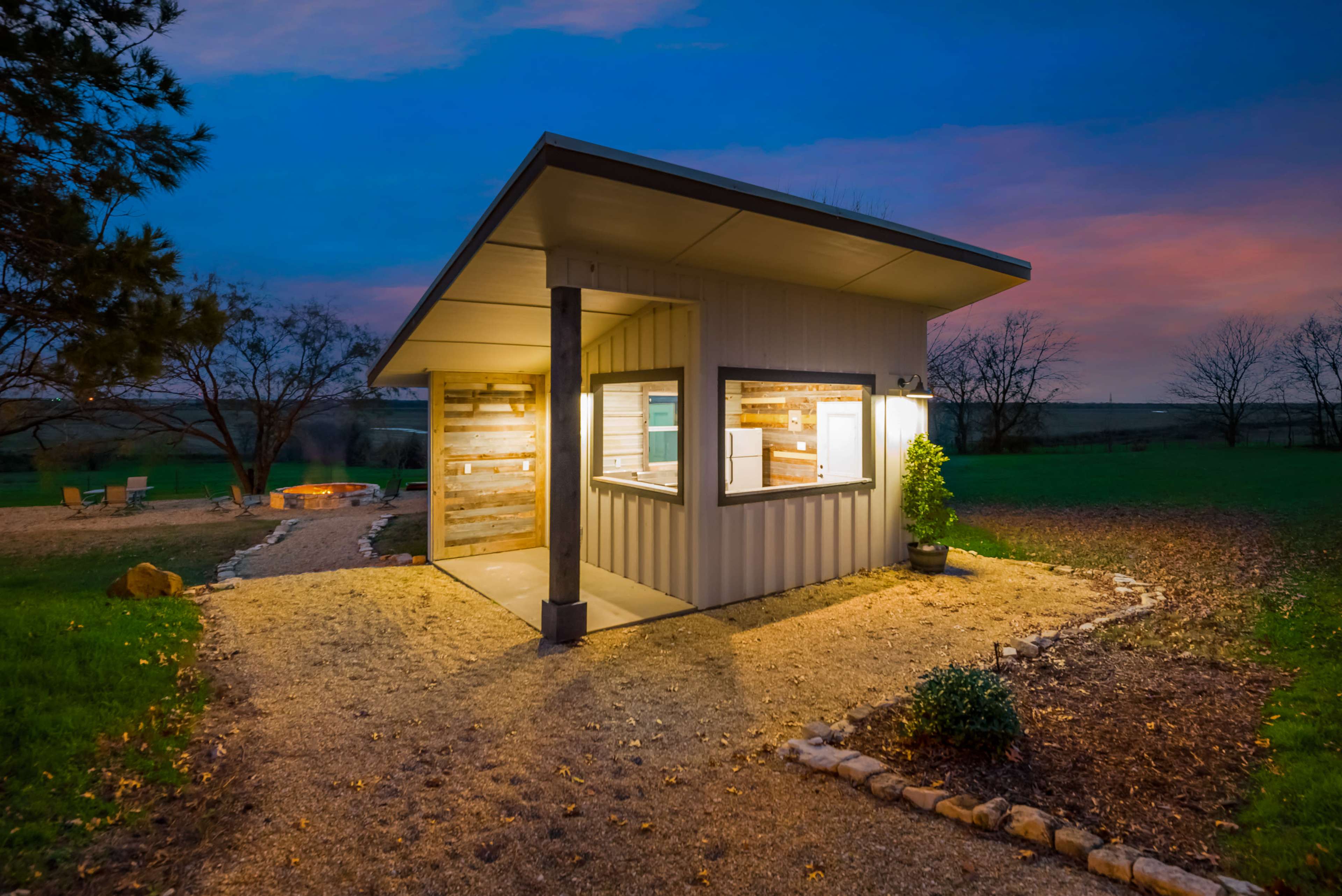 A modern cabin with large windows is illuminated at dusk, situated on a gravel path surrounded by trees and outdoor seating.