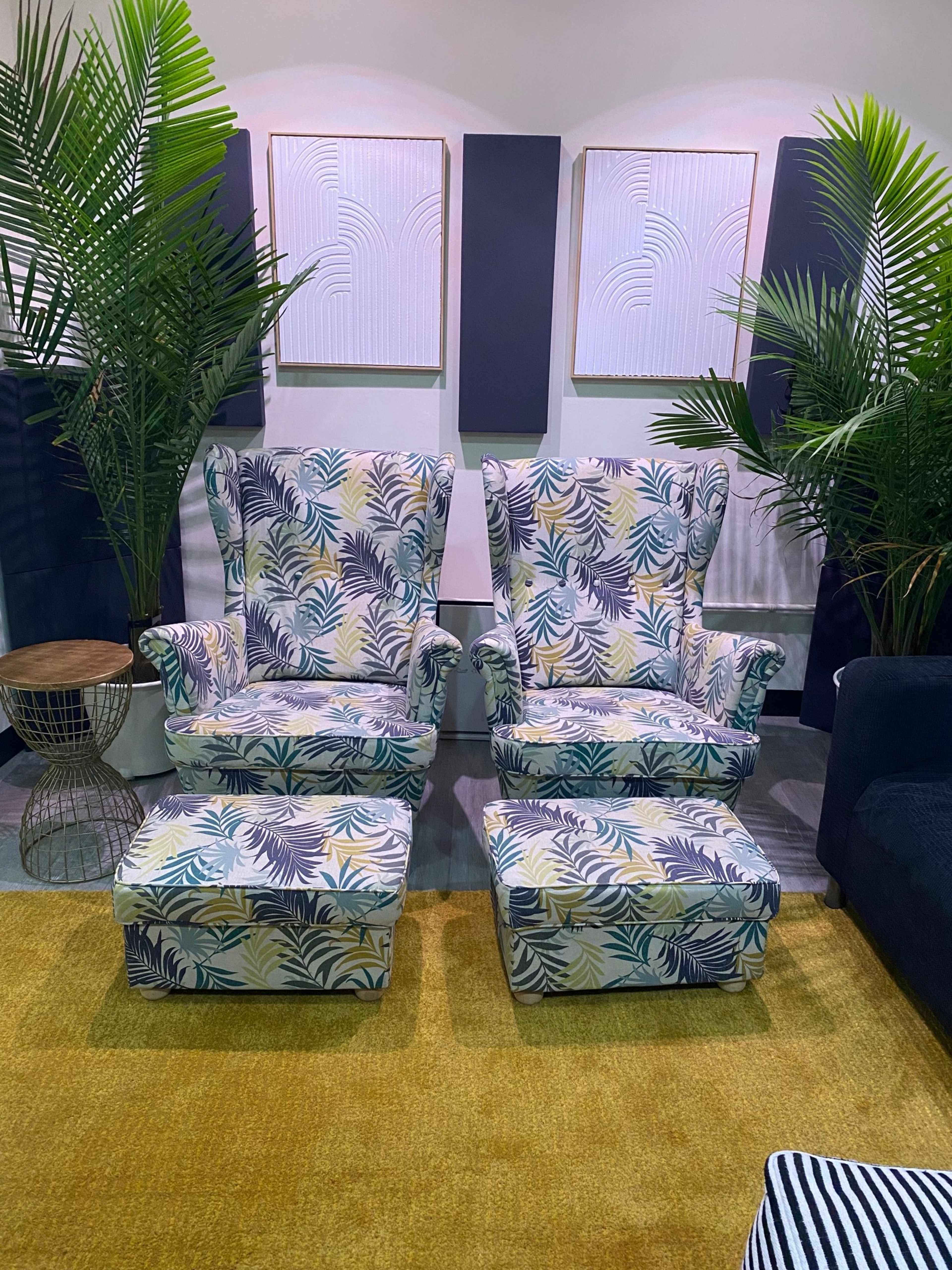 Two patterned armchairs with matching ottomans, placed between leafy plants and decorated wall art in a modern interior setting.