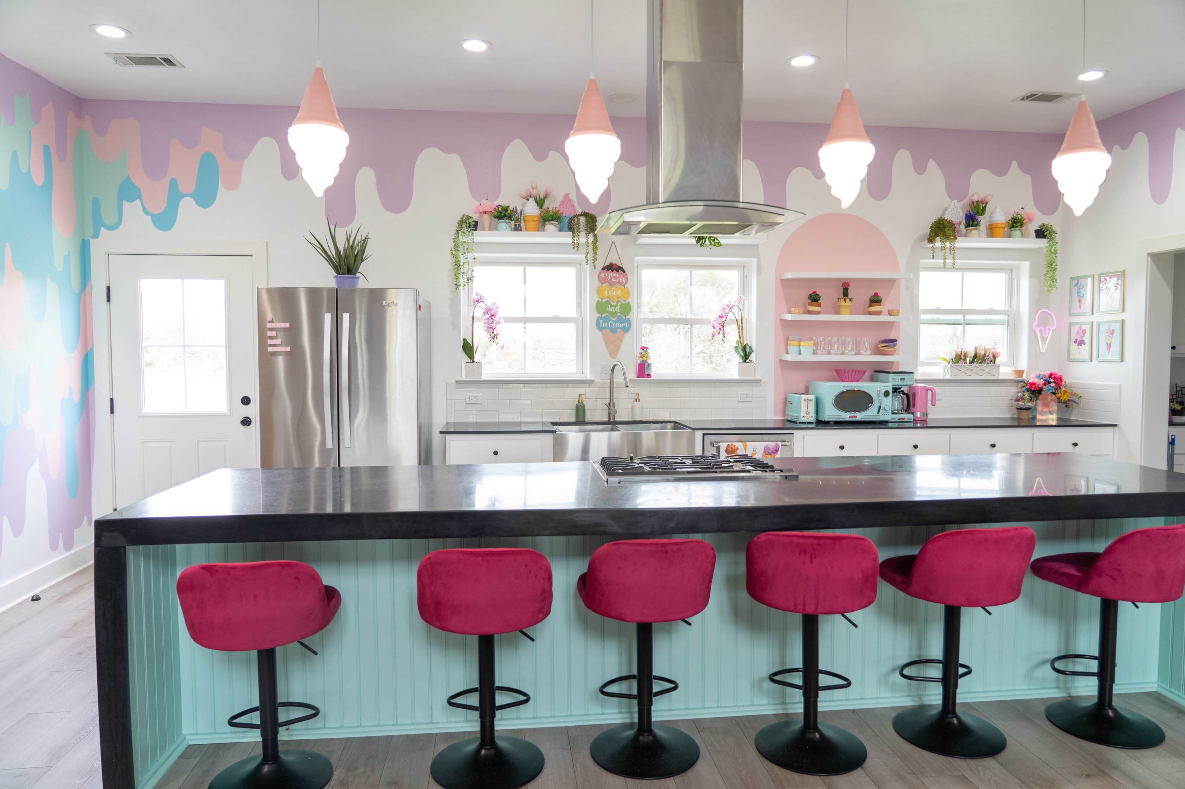 A colorful kitchen features a large island with pink bar stools, stainless steel appliances, and a backdrop of pastel-painted walls.