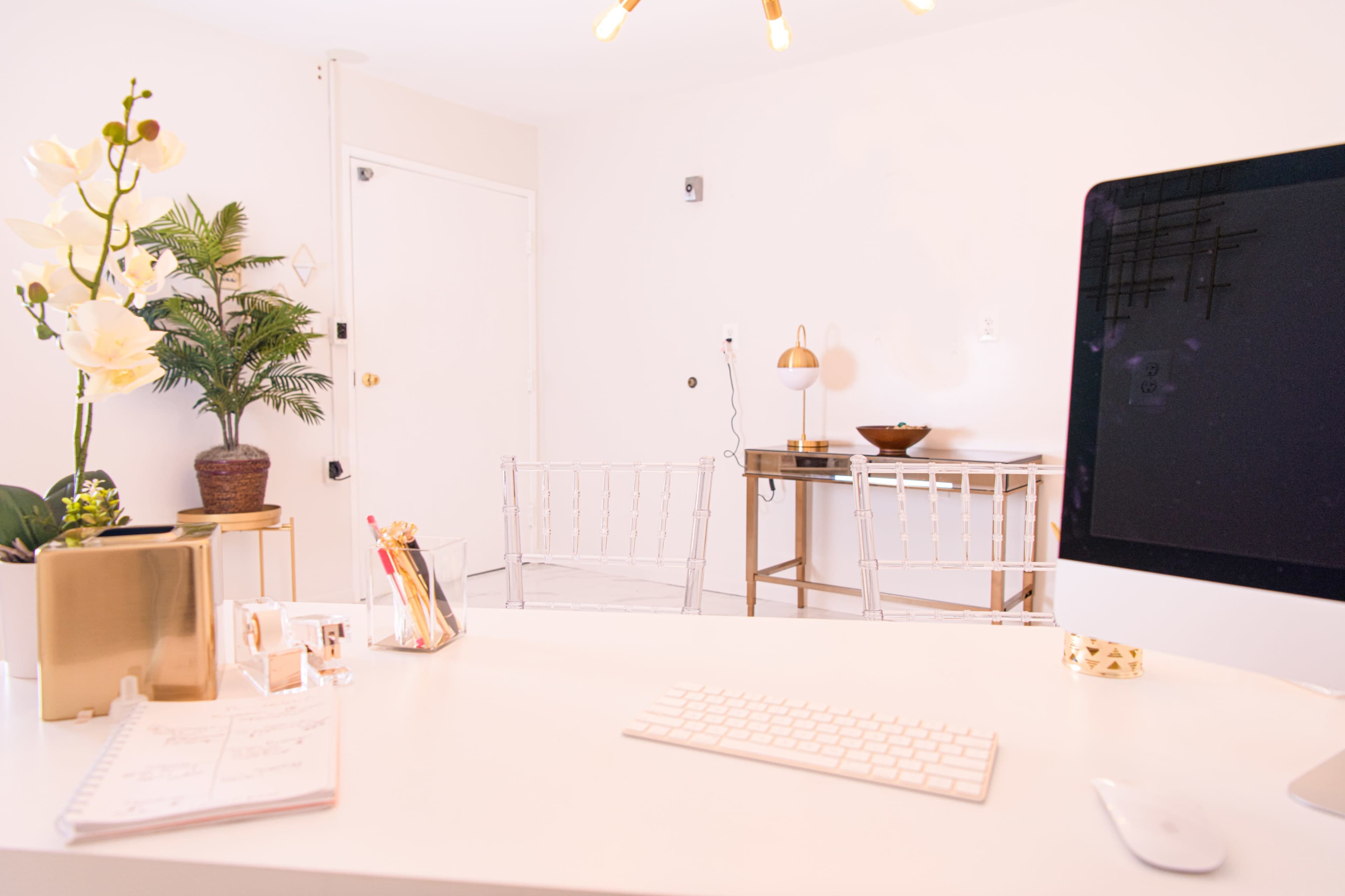 An office space features a desk with a computer, stationery, and a small plant, with two transparent chairs and a decorative table in the background.