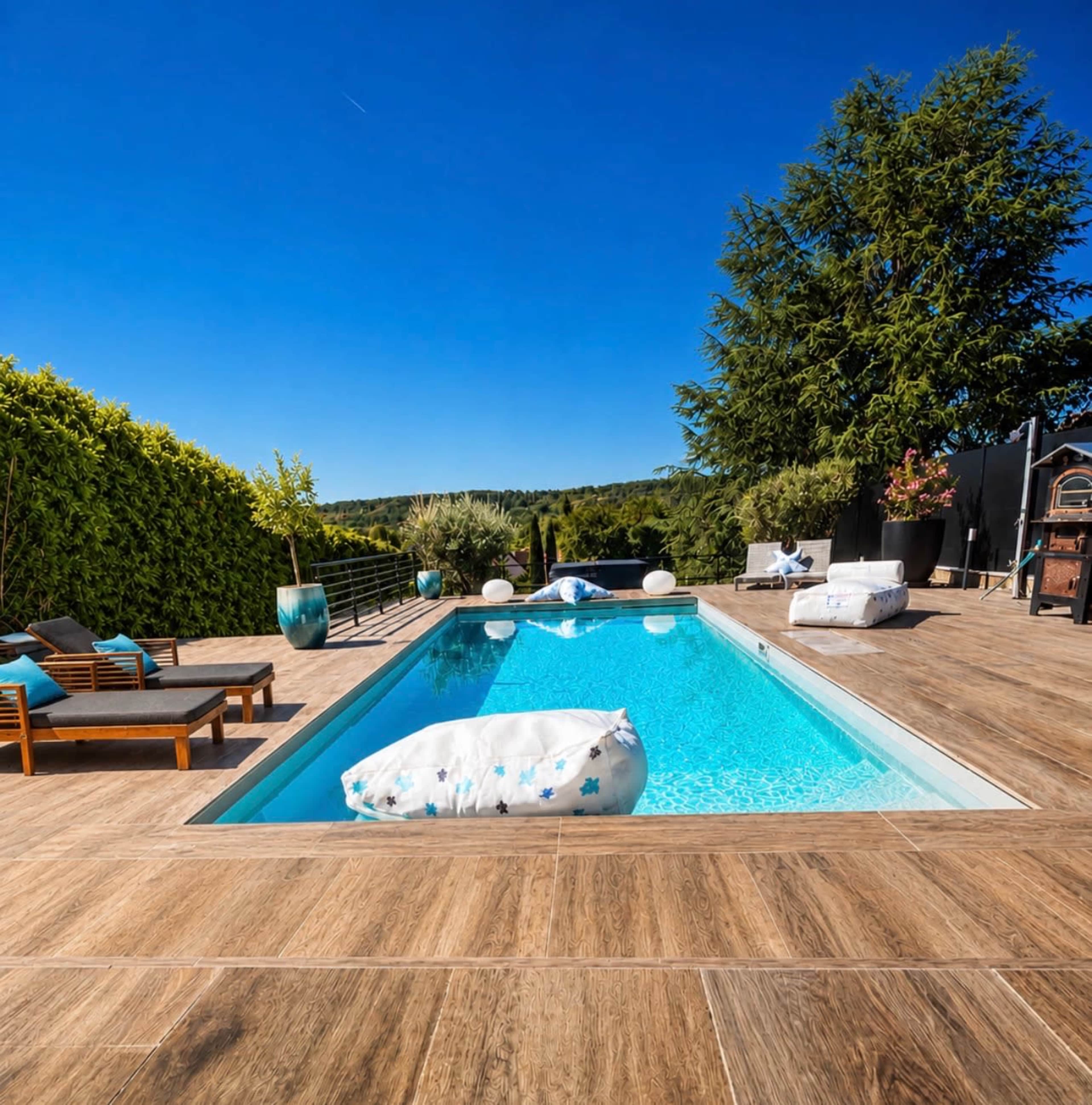 A modern outdoor pool surrounded by wooden decking and lounge furniture, with greenery and clear blue skies in the background.