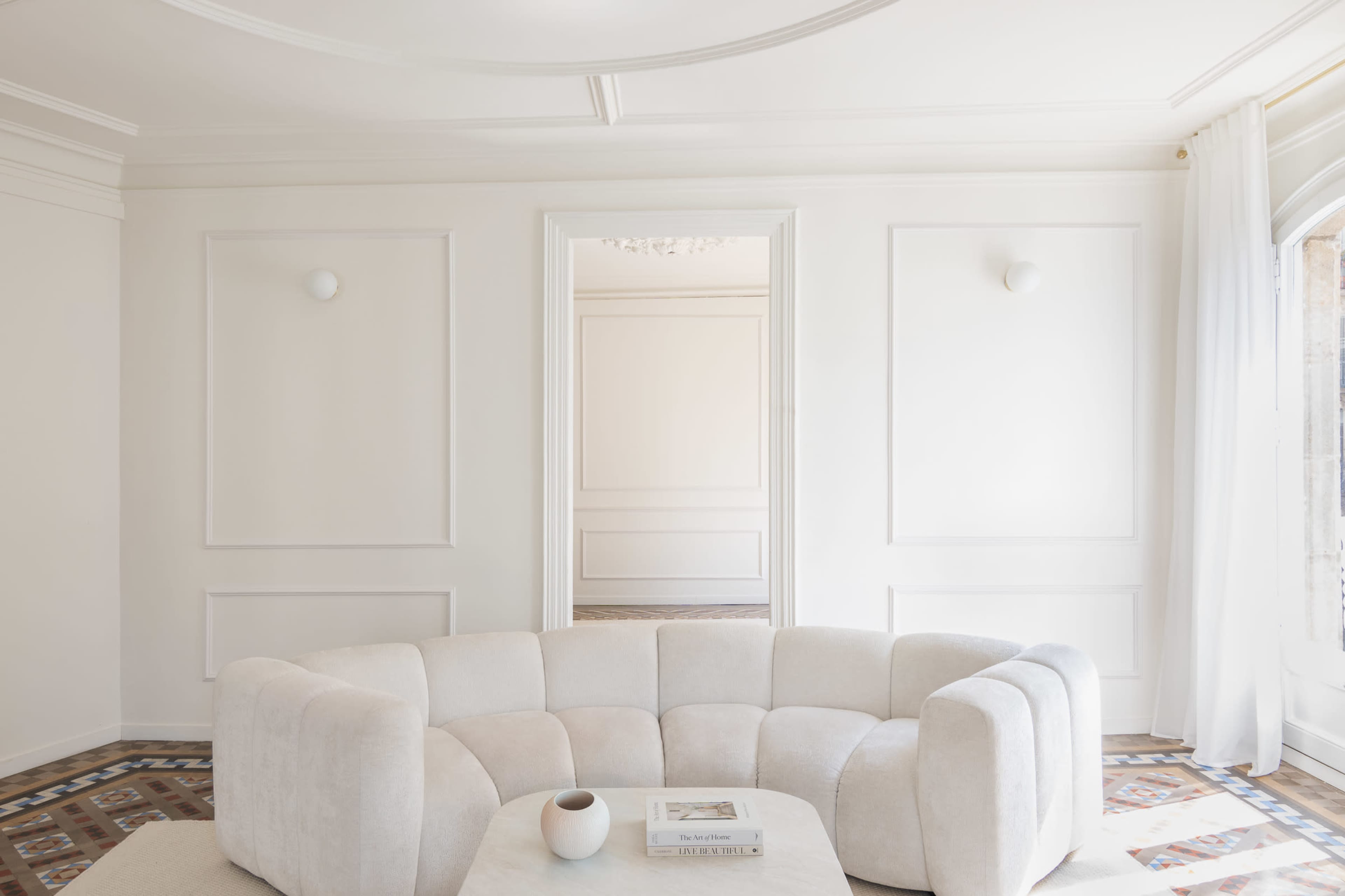 The image shows a minimalist living space with a curved sofa and a coffee table, featuring white walls and patterned flooring.