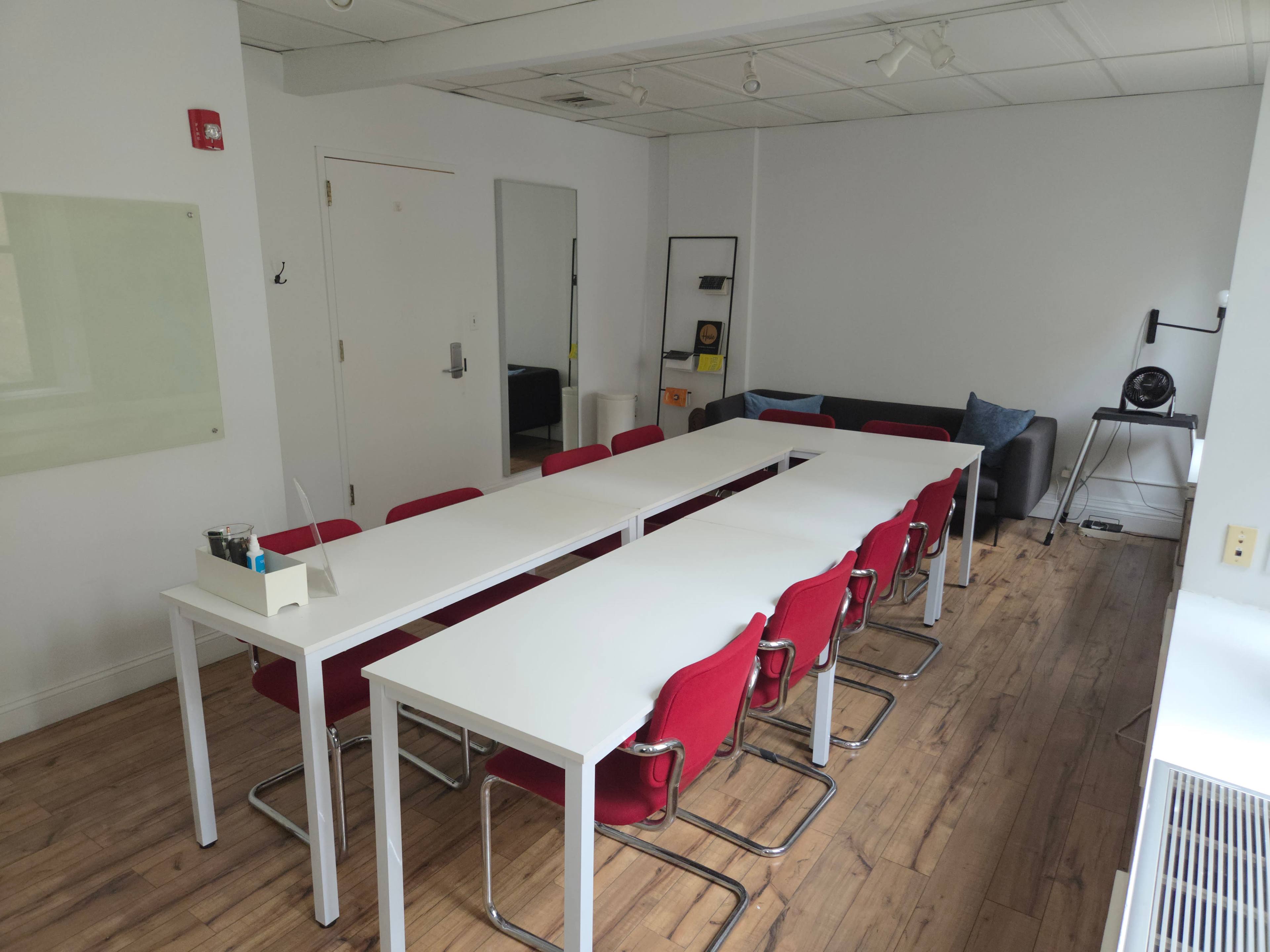A bright meeting room features a long white table surrounded by red chairs and has a sofa against the wall.