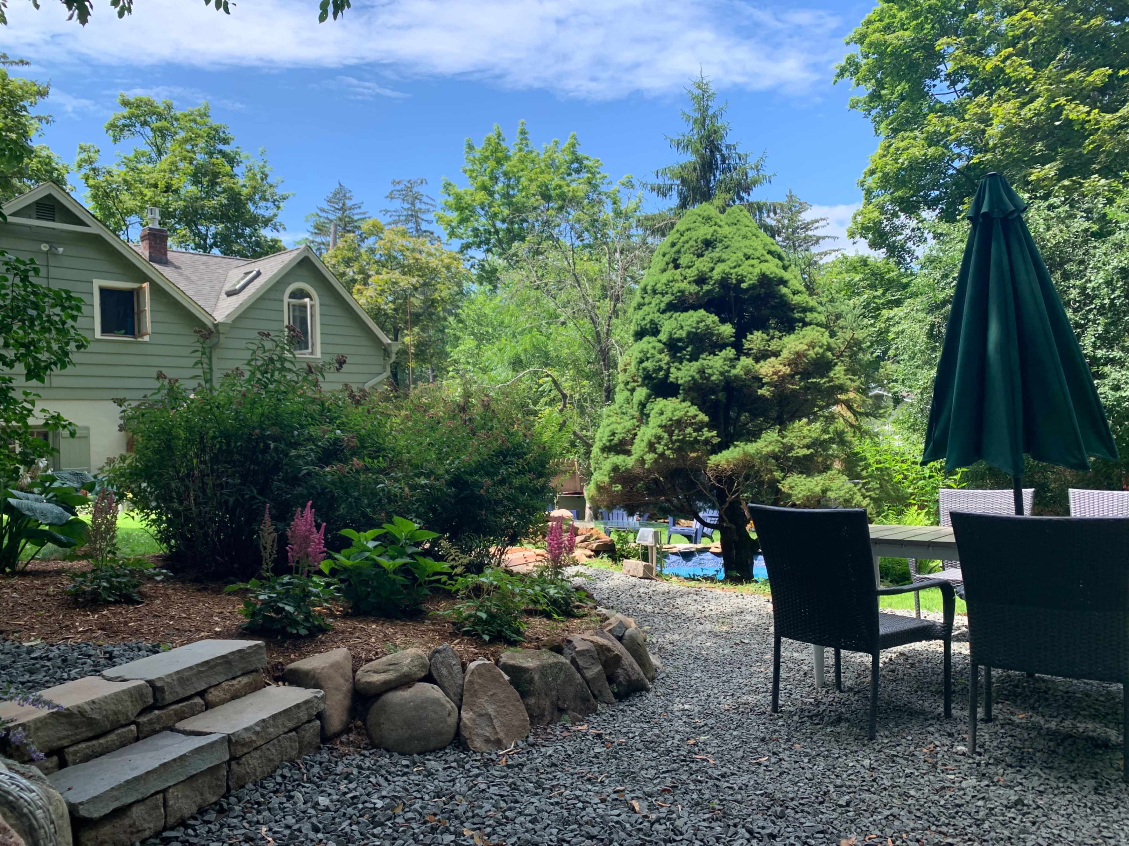A stone path leads through a garden area with greenery and a patio set beneath a large umbrella, alongside a house in the background.